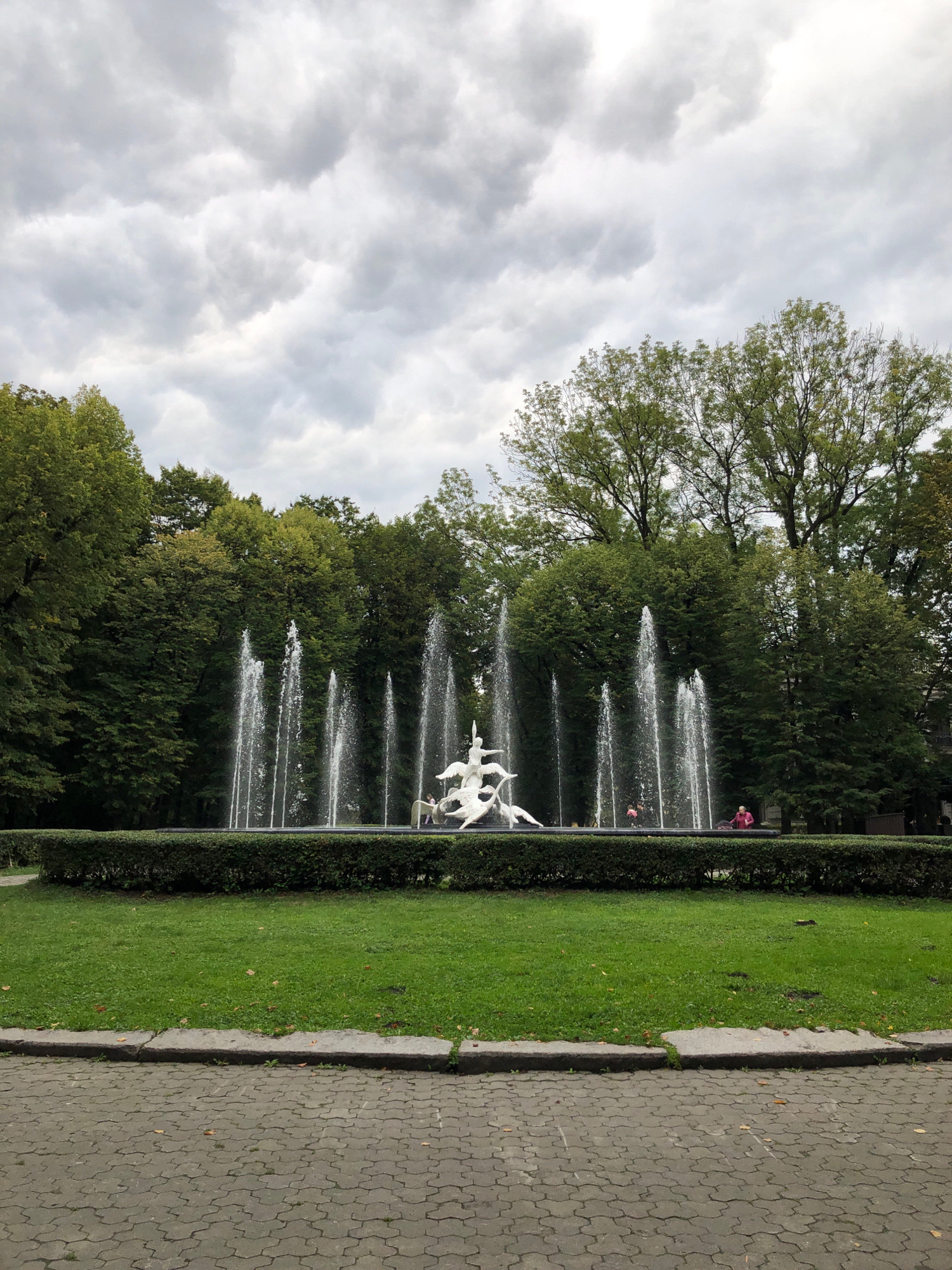 The image depicts a public fountain located in Lviv, Ukraine, specifically in the Ivan Franko Park. The fountain features a central white sculpture of figures in dynamic poses, surrounded by several jets of water reaching upwards. The fountain is situated in a rectangular basin, bordered by a manicured hedge. Behind the hedge, a lush green lawn is visible, with scattered autumn leaves. The background consists of a dense line of tall trees with full green foliage, suggesting a park setting. The sky above is overcast with heavy, grey clouds. In the foreground, a paved walkway made of interlocking grey pavers leads towards the park, with a stone border separating it from the grassy area. Two individuals are faintly visible near the fountain; one appears to be sitting on the edge of the fountain's basin, while another is standing. The overall atmosphere is subdued, likely due to the cloudy weather.