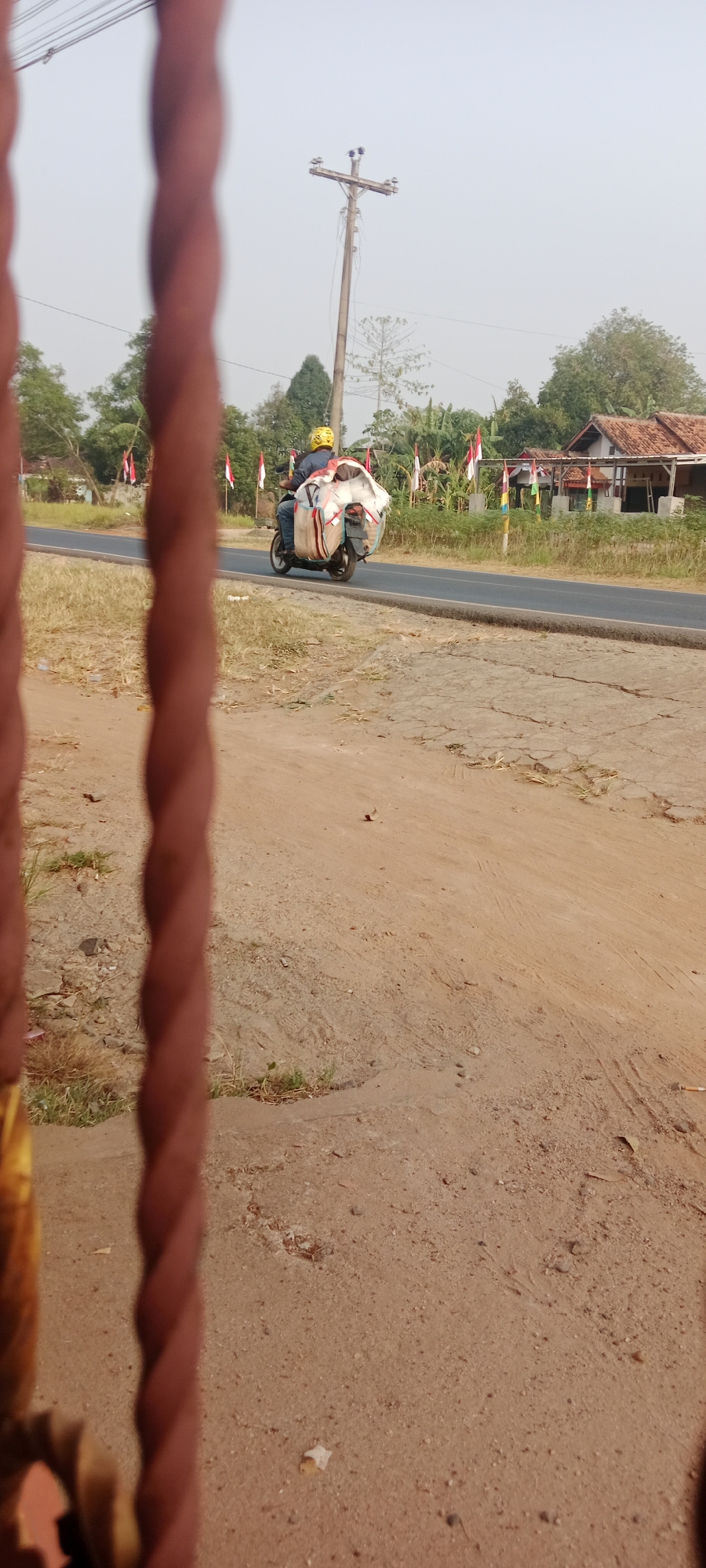 In the city of Gondang Rejo, Indonesia, a person wearing a yellow helmet rides a motorcycle on a paved road. The motorcycle is loaded with a large, white, fabric-wrapped object, possibly a mattress or bulky packaging, secured to the rear. The rider is wearing a dark shirt and blue jeans. The road runs through a rural area with dry, grassy fields on either side. In the background, there are trees, a utility pole with wires, and a building with a tiled roof. Several small Indonesian flags are also visible along the roadside, suggesting a local event or celebration. The scene is captured through a blurred foreground of what appears to be a fence or railing, creating a sense of depth and a slightly obstructed view. The sky is overcast, and the lighting suggests it is daytime.