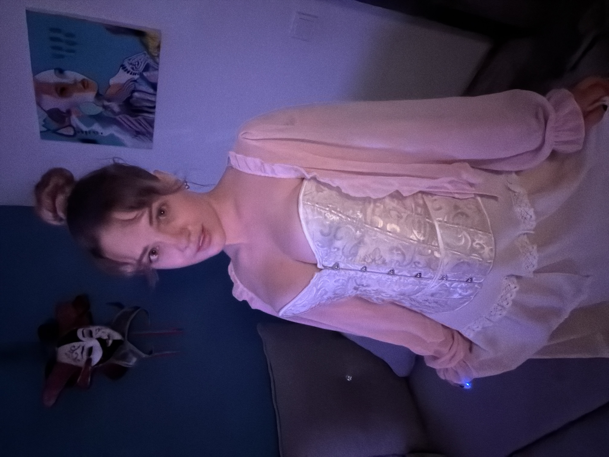 A young woman with dark hair tied in a bun and bangs is positioned diagonally across the frame, looking directly at the viewer with a neutral expression. She wears a pale pink, long-sleeved top with ruffled cuffs, over which she has a light-colored, possibly white or silver-grey, ornate corset featuring textured or embroidered patterns and several visible hook-and-eye closures down the front.
The scene is indoors, characterized by dim lighting with a strong blue and purple cast. Behind her head, a framed picture or poster with a stylized portrait in blue and purple tones hangs on the wall. To her left, on a dark blue cushion or sofa, lies a decorative mask, predominantly black, white, and red. The woman is seated or reclining on a dark gray upholstered piece of furniture. A small, bright blue light emanates from an object near her right hand. No visible text is present in the image. The setting is an indoor space, within the contextual location of Большая Шильна, Russia.
