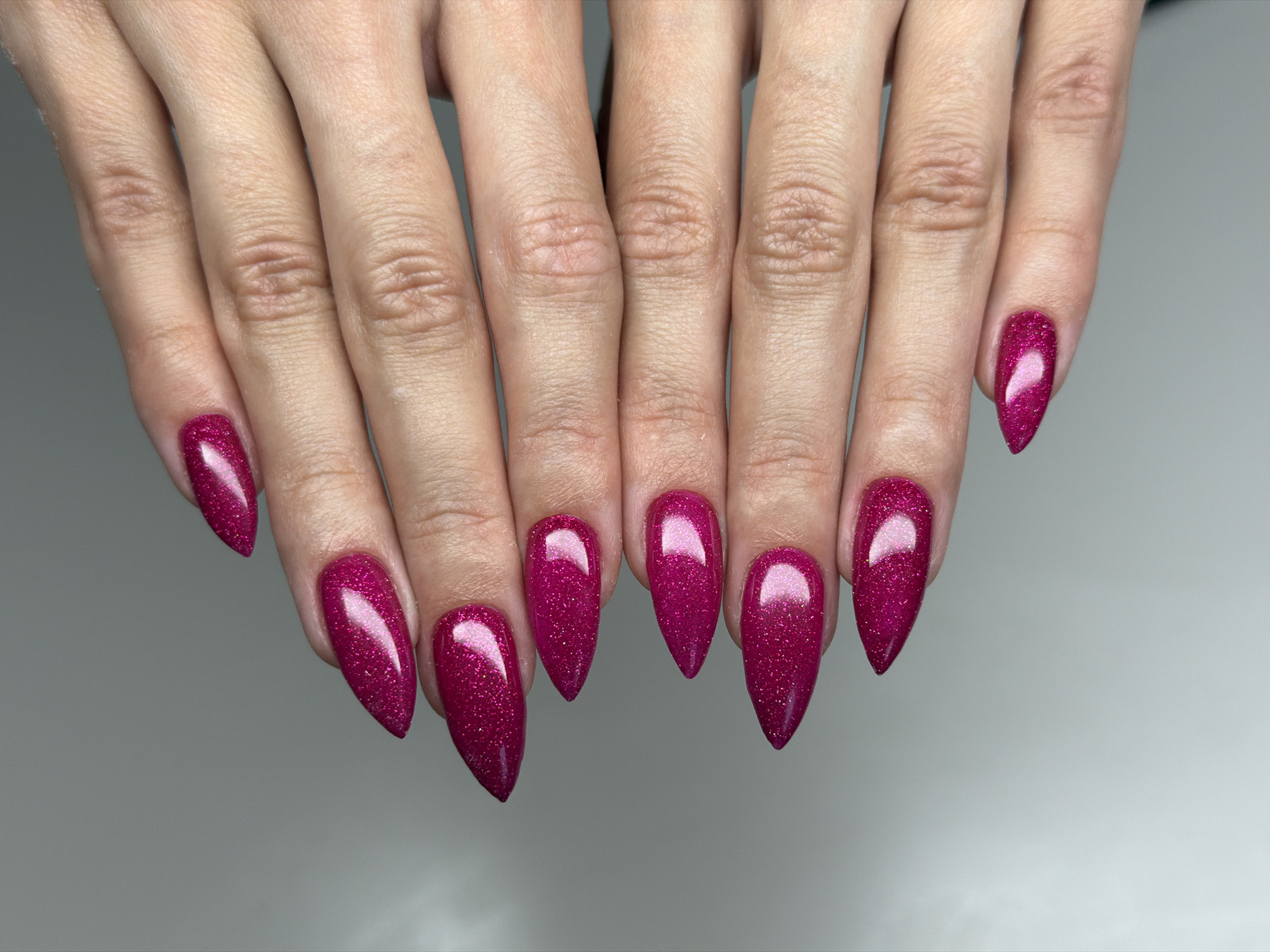 The image displays a pair of hands, presented with fingers extended and slightly spread, palms down, against a plain, light grey background. The central focus is on the ten fingernails, which are uniformly long and shaped into a sharp almond or stiletto style. Each nail is coated in a vibrant, glittery deep magenta or fuchsia polish, with visible sparkle and prominent bright reflections on their surfaces.
The skin on the hands and fingers is light to medium in tone, showing natural creases, wrinkles, and some dryness, particularly noticeable around the knuckles and finger joints. The lighting is soft and even, suggesting an indoor, controlled setting, likely a nail salon or studio. The overall impression is one of a detailed beauty shot showcasing a manicure. No specific activity is occurring beyond the posing of the hands for display. There is no visible text in the image. This scene is located in Ramat Gan, Israel.