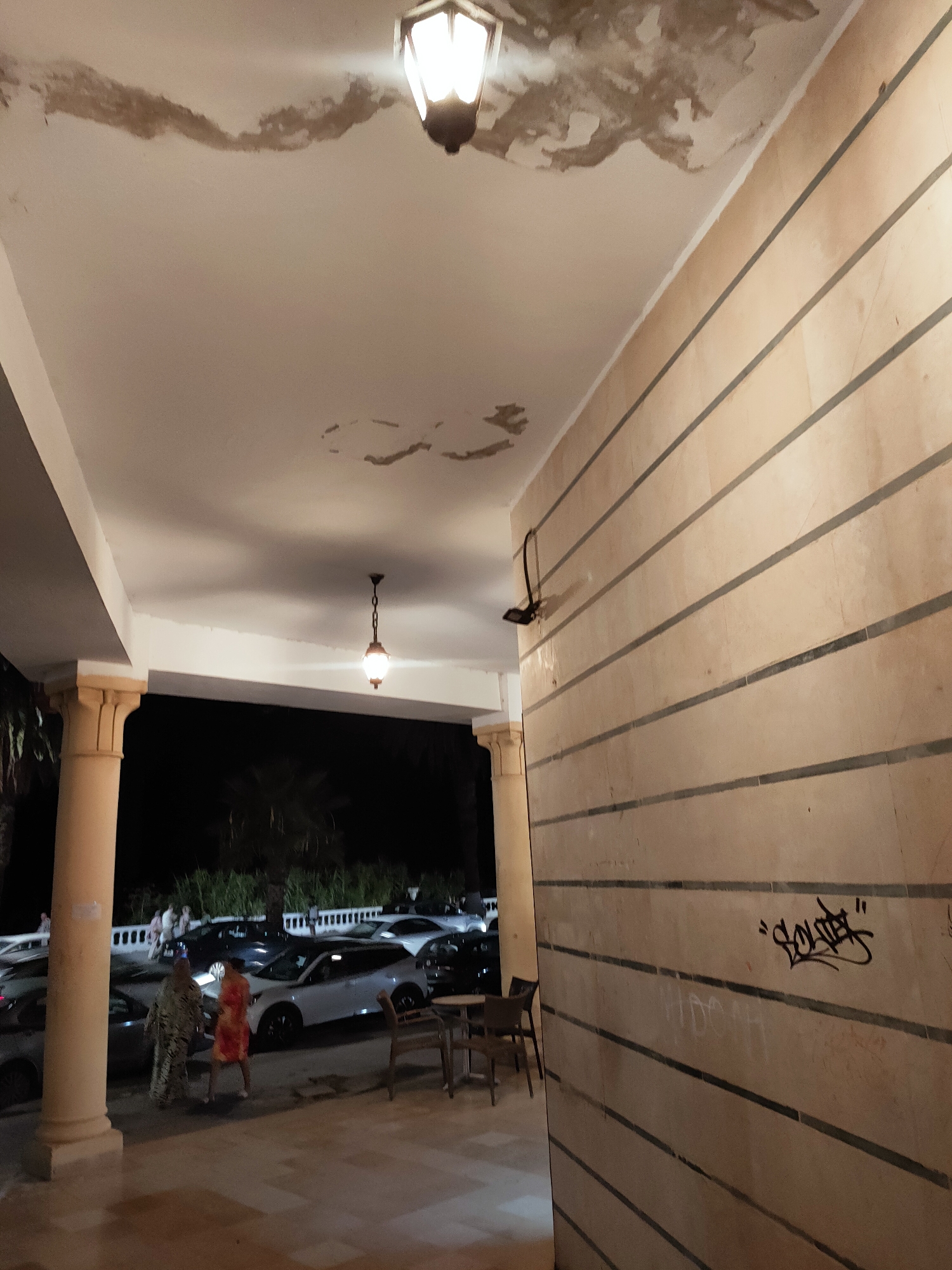 The image captures a section of an outdoor covered walkway at night. The ceiling of the walkway shows signs of water damage, with peeling paint evident. Two light fixtures are visible: one is a modern, square lamp attached to the wall, and another is a more traditional hanging lamp with a decorative chain. The walkway wall is covered in horizontally striped, light-colored paneling. Graffiti in black ink is present on this wall.
In the background, a dimly lit street scene unfolds with several parked cars, including what appear to be white sedans and SUVs. A few palm trees are visible, suggesting a warm climate. Two individuals, appearing to be women, are walking on the pavement in the vicinity of the parked cars. One is wearing a patterned dress, and the other is in a brightly colored orange and red dress. A small table and chairs are set up near the walkway, hinting at an outdoor seating area for a cafe or restaurant.
Based on the presence of Arabic script in the graffiti and the architectural style, the location is likely in the city of Tunis, Tunisia. The overall atmosphere is one of an urban setting at night, with elements of both decay and daily life.