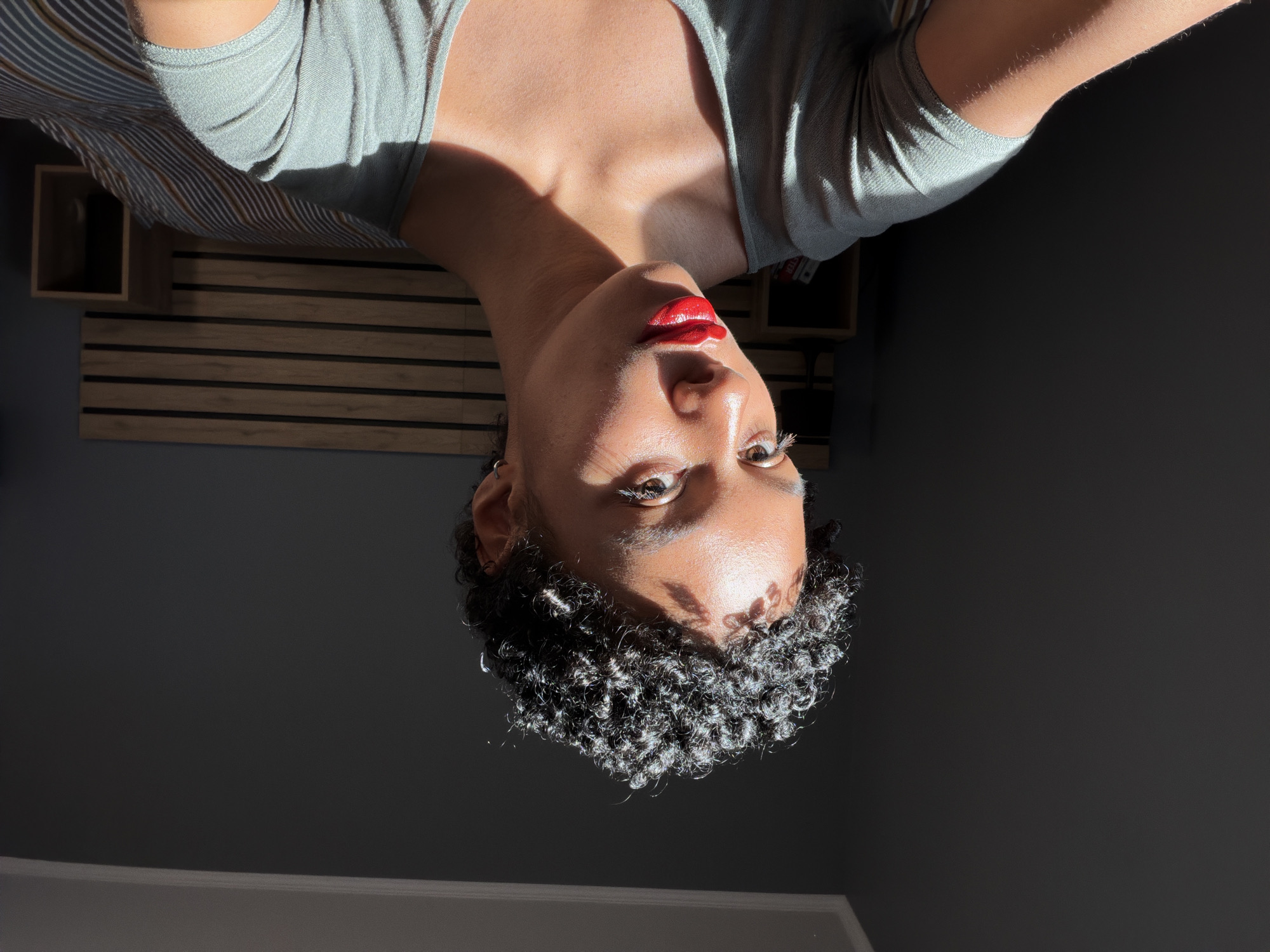 A woman with short, tightly curled dark hair, wearing a grey top and bright red lipstick, is seen in a close-up shot with the camera positioned below her looking upwards. The background consists of a dark wall and a partially visible wooden slat wall decoration. The setting appears to be indoors with strong, direct sunlight illuminating portions of her face and hair. There is no evidence to suggest this image was taken in Champs-sur-Marne, France.