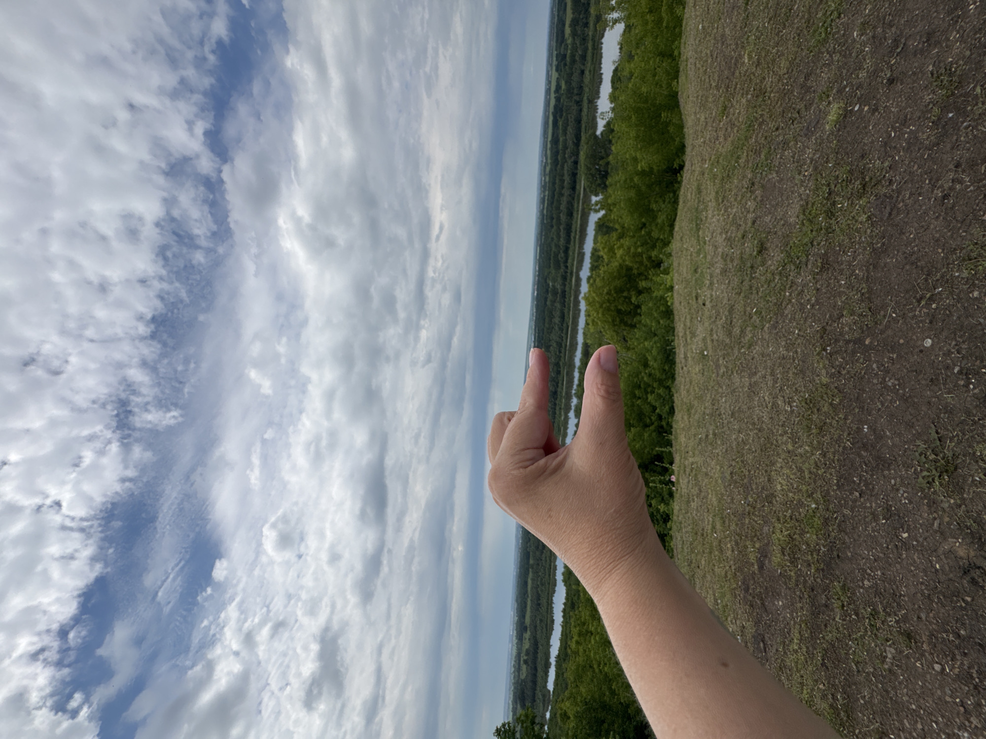 A first-person perspective shot captures a hand gesturing "small" against a panoramic view of the city of Ufa, Russia. The hand, positioned in the lower left quadrant, is illuminated and in focus, with fingers pinched together to indicate a small size. Beyond the hand, a lush, green landscape unfolds, featuring a meandering river or body of water reflecting the cloudy sky. Trees and vegetation dominate the midground, transitioning to the distant urban landscape of Ufa under a dramatic, cloud-filled sky. The foreground consists of a dirt and grass embankment.