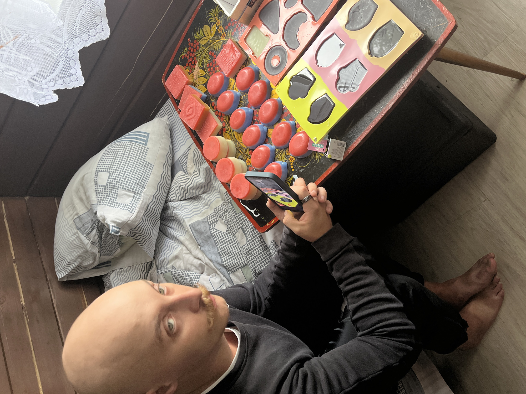 A bald man with a mustache is sitting on a bed, holding a smartphone. He is wearing a black sweatshirt and black pants. The bed has a pillow with a patterned cover. A wooden table with a black surface and red trim is next to the bed, covered with various soaps. The soaps are in different shapes and colors, including red, pink, and blue. Some of the soaps are in molds. A white lace curtain is visible in the background near a window. The floor is made of light-colored wooden planks. The overall impression is an indoor setting, possibly a room in a house. The location is specified as Divnogorsk, Russia, though this is not visually evident from the image.