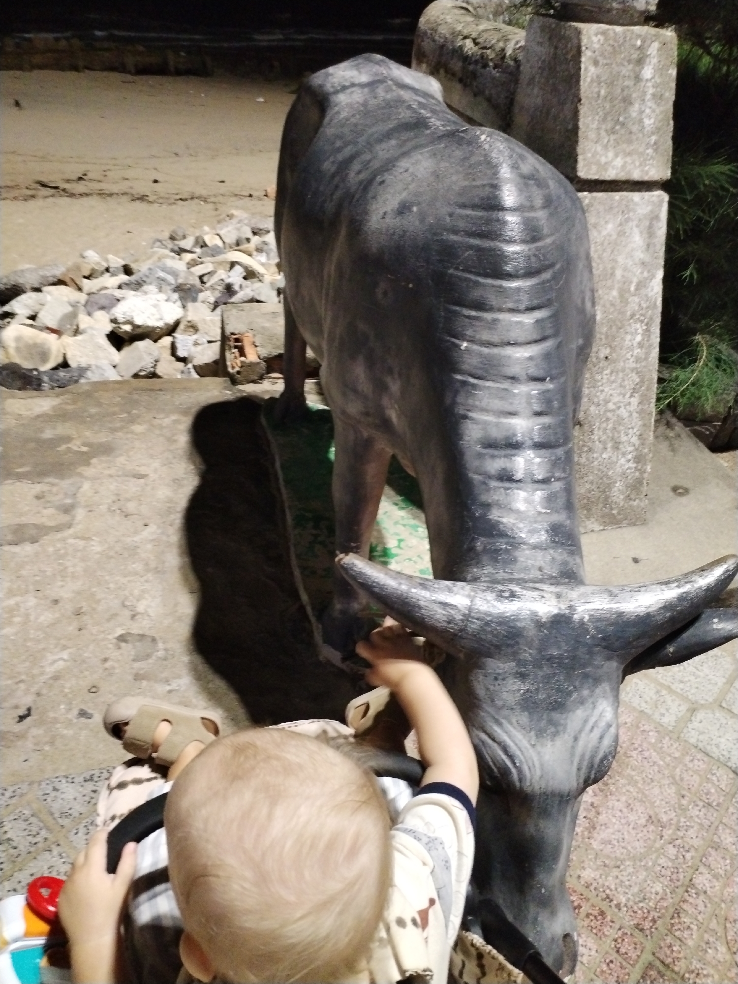 A high-angle, full shot taken in Nha Trang Ward, Vietnam shows a toddler in a stroller interacting with a statue of a water buffalo. The toddler, with a light-colored head of hair, is seen from the back. The child's right arm is raised toward the buffalo's horn, indicating interaction. The toddler is seated in a stroller with visible black handles and is wearing a cream and light-colored patterned shirt with a blue-trimmed sleeve. A light brown sandal is visible on the toddler's left foot. The water buffalo statue is predominantly dark grey and appears to be made of concrete or a similar material. It is positioned next to a concrete pillar with a rough, weathered texture. The ground is a mix of concrete and patterned tiling. In the background, there is a pile of rocks and vegetation. The lighting suggests it is either dusk or dawn.