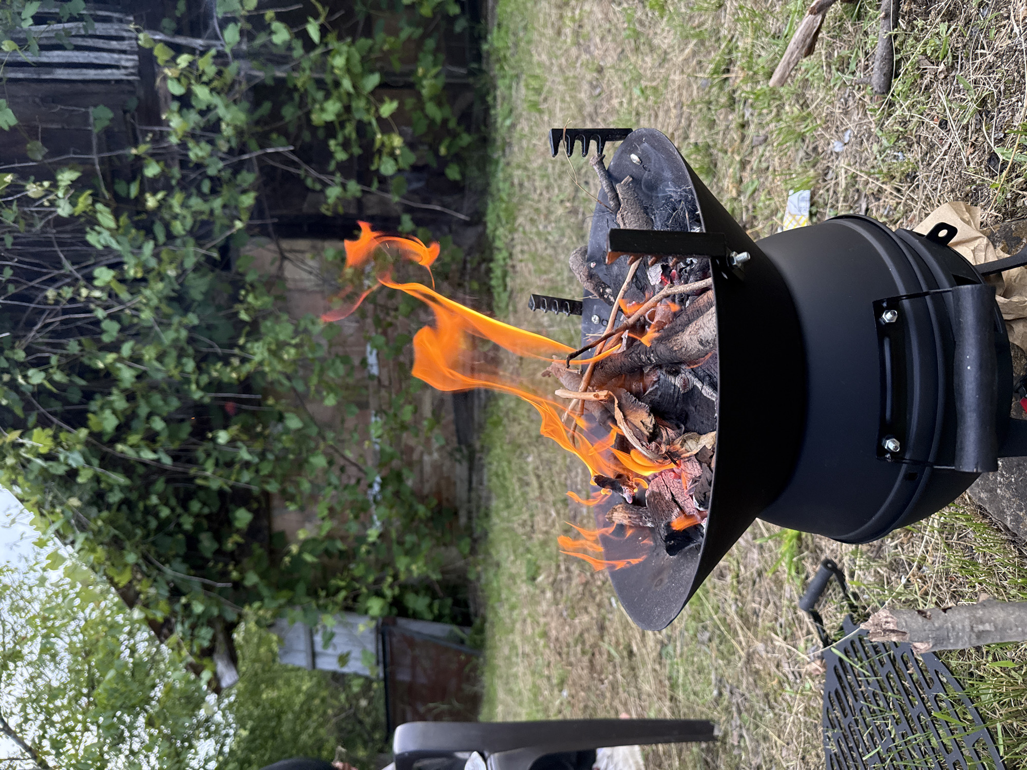 This image, taken in Solari, Bulgaria, showcases an outdoor scene centered around a black, barrel-shaped metal fire pit or grill. A vibrant fire burns actively within the pit, producing prominent orange flames and some smoke. Inside, a mixture of burning wood logs, kindling, and glowing embers is visible. The fire pit features black metal handles and support structures.
The surrounding environment consists of patchy green grass and areas of dry soil. To the left, dense green foliage, likely vines or bushes, grows over and around an old wooden structure, possibly a fence or part of a building. Further in the background, another wooden building is partially obscured by the greenery. On the ground near the fire pit, a black metal cooking grate and a separate black handle are placed. A crumpled brown paper bag and a small, indistinct white wrapper are also visible on the ground.
The scene is brightly lit by natural light, indicating it is daytime, likely midday or early afternoon, under clear weather conditions. No individuals are present, but the active fire suggests an ongoing outdoor activity, possibly preparing for cooking or simply enjoying a campfire. No discernible text is visible anywhere in the image.