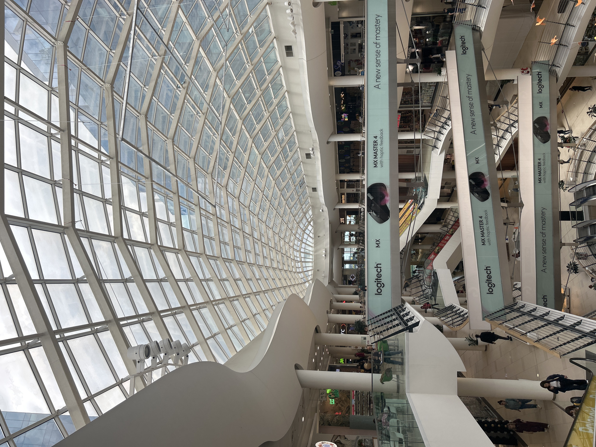 Here is a description of the image:
The image captures the interior of a multi-level shopping mall, notable for its vaulted glass ceiling supported by a grid-like structure. Several large white supporting structures add to the airy feel of the place. Banners advertising Logitech products are suspended from the ceiling. Escalators and retail spaces are visible in the background. People can be seen walking and interacting within the mall.