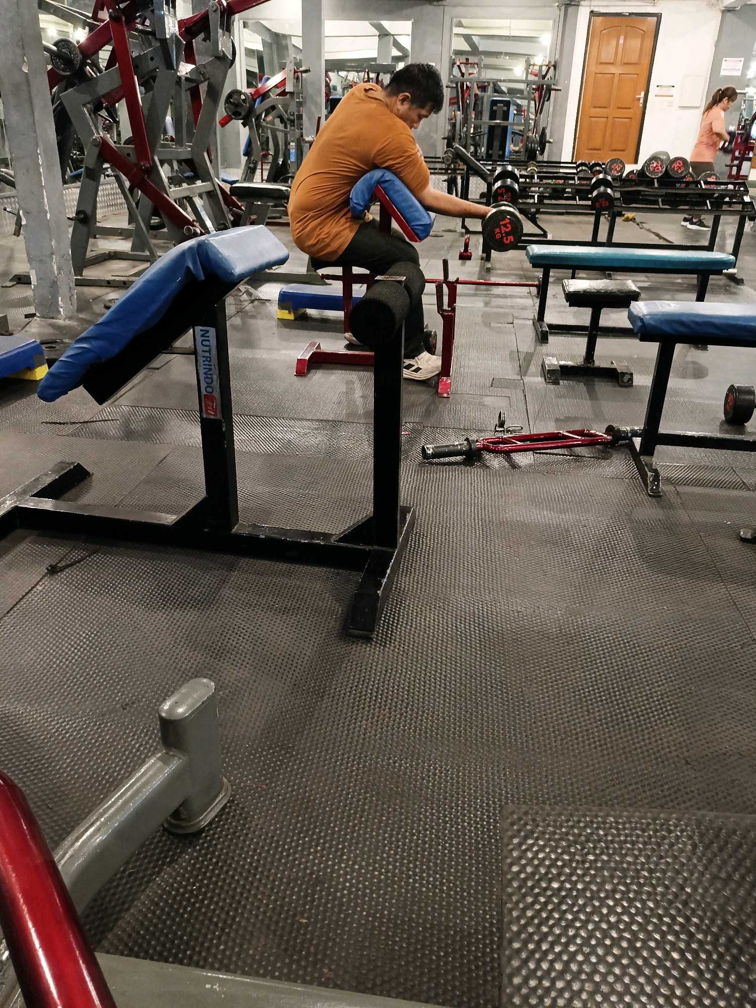 An overhead medium shot shows a man working out in a gym. The man is wearing an orange t-shirt and black pants and is sitting on a preacher curl bench with a dumbbell in his right hand. He is performing a bicep curl. In the background, there are gym equipment, including weight machines and benches, and a woman is walking by. The gym floor is black rubber matting. The lighting is dim. The man is the central focus of the image. There is a red and silver EZ curl bar on the floor near the preacher curl bench.
