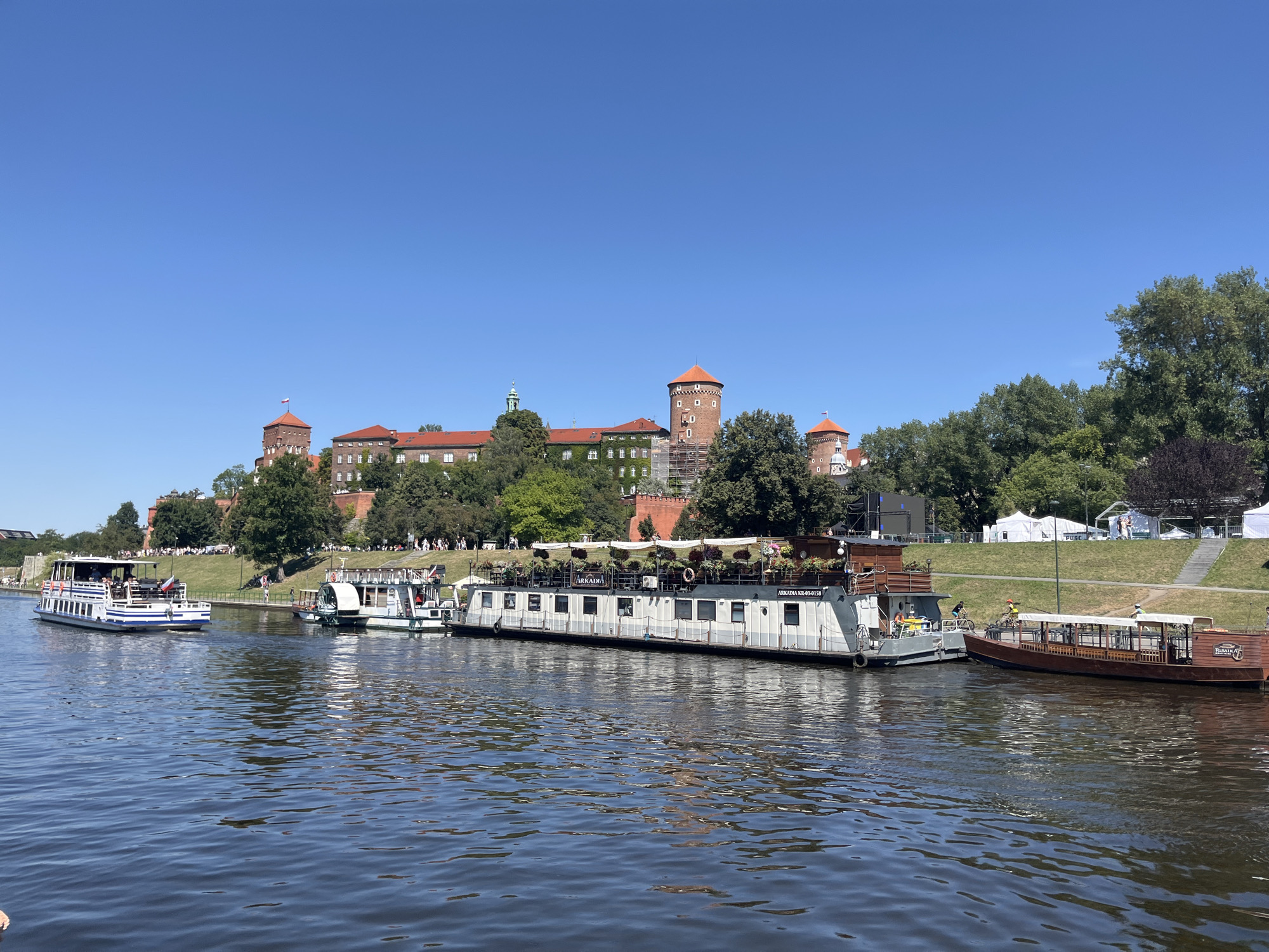 The image captures a wide view of the Vistula River in Krakow, Poland, with Wawel Castle prominently situated on the embankment in the background. Several tour boats are docked and navigating the river. On the left, a white and blue passenger boat is moving away from the camera. To its right, a larger, white and grey boat with an outdoor seating area and the name "ARKADIA" visible on its side is docked. Further to the right, a smaller, reddish-brown wooden boat with a covered seating area is also docked. The castle complex features red-tiled roofs and several towers, one of which displays a flag. Lush green trees line the embankment, and the sky is clear and blue. A grassy slope with a path and scattered individuals is visible between the river and the castle.
