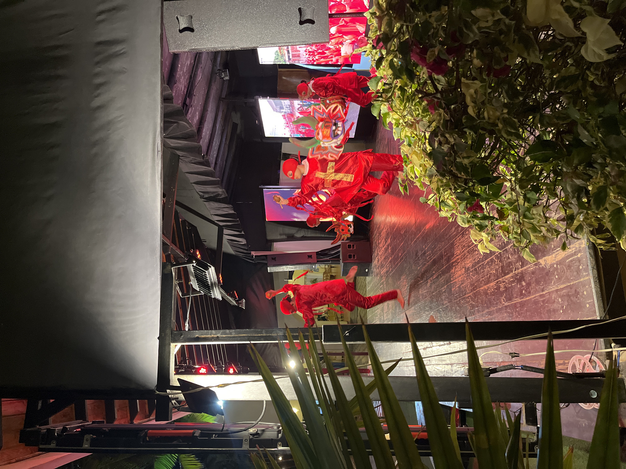 In Porlamar, Venezuela, a stage performance is captured at night, likely within an outdoor or semi-outdoor venue. Several performers, dressed in striking red costumes that include matching head coverings, are actively engaged on a wooden stage. Two individuals in the foreground are holding and manipulating a large, colorful dragon prop, which features prominent green horns and a fierce expression. The costumes are adorned with large gold crosses, suggesting a possible religious or symbolic theme for the performance.
Behind the performers, large LED screens display vibrant images, contributing to the dynamic visual environment. The stage is illuminated by a mix of red and white lights, casting a warm glow and creating dramatic shadows. The surrounding environment appears to be a venue with structures overhead, including what seems to be a black fabric canopy and lighting rigs. To the left and right foreground, lush green foliage partially obstructs the view, indicating the presence of plants or landscaping around the performance area. The overall scene suggests a lively cultural event or entertainment performance taking place in the city.