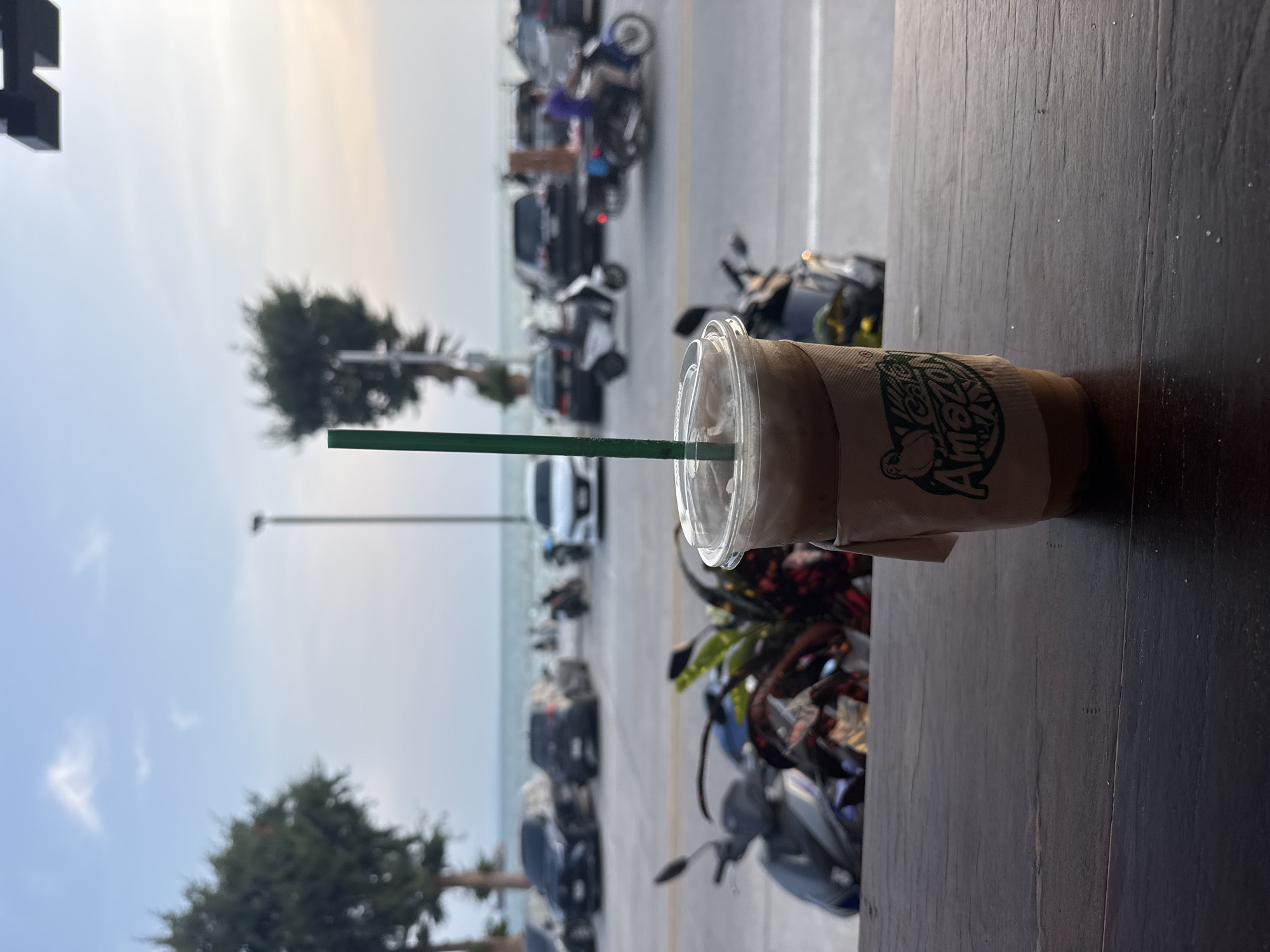 The image, oriented on its side, presents an iced coffee drink resting on a dark wooden surface, overlooking a street scene and a distant body of water.
In the foreground, a clear plastic cup holds an iced coffee, topped with a lid and a green straw. A brown paper sleeve wraps around the cup, prominently displaying the green "CAFE AMAZON" logo, which includes a stylized 'A' and a graphic resembling a bird or parrot. The coffee sits on a dark, heavily textured wooden table or counter.
Beyond the table, a street is visible, albeit out of focus, filled with numerous parked and moving vehicles, primarily motorcycles and some cars. A yellow line marks the edge of the road. Further in the background, a hazy horizon separates the land from a calm, blue-gray expanse of sea or a large body of water. The sky above is bright and mostly clear, with faint, wispy clouds, suggesting daytime.
Two green, leafy trees are visible in the left mid-ground, and a thin, tall pole extends upwards into the sky. The overall scene depicts a casual outdoor setting, likely a cafe or eatery, situated alongside a road near a coast. The prevalence of motorcycles and the seaside view align with a typical streetscape in Pattaya City, Thailand. No individuals are clearly visible in the frame.