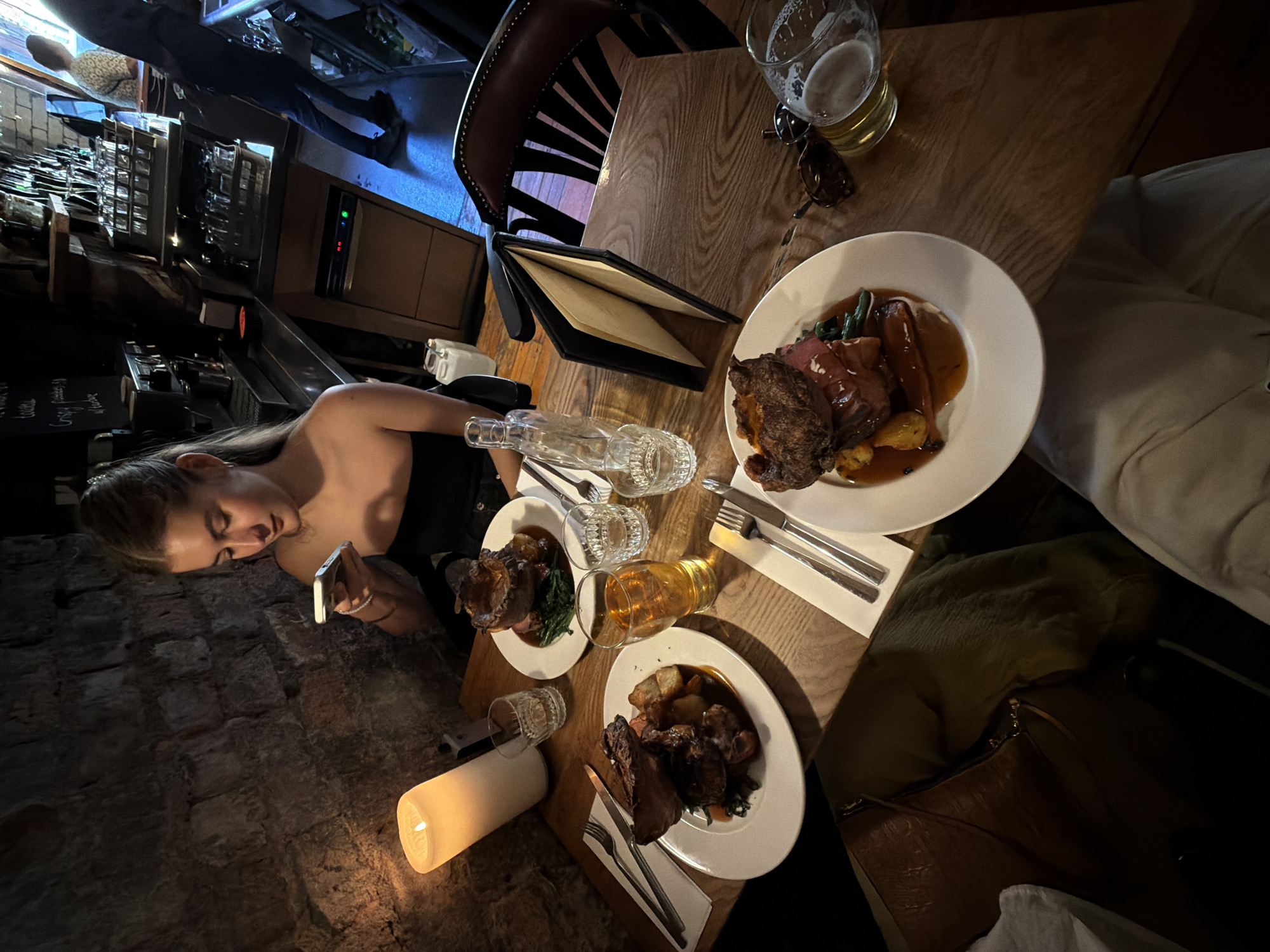 The image shows a dimly lit interior, possibly a restaurant or pub, with a rustic aesthetic featuring a brick wall. A woman is seated at a wooden table, using a mobile phone. The table is set for a meal, with at least two plates of food that appear to be roasts with gravy and vegetables. There is also a beer glass, a clear bottle, cutlery, and a lit candle providing ambient light. A menu is open on the table, and a handbag is placed nearby. The location is unspecified; the city is Unknown, Unknown.