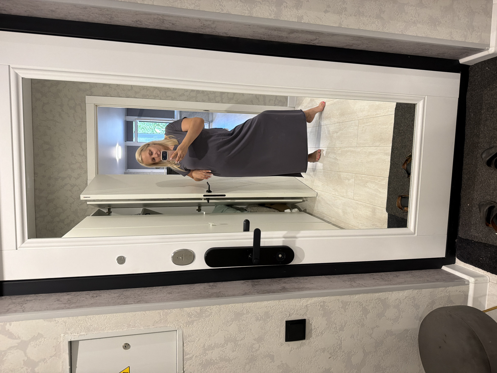 An adult Caucasian female is standing in a doorway, taking a mirror selfie. She is wearing a dark gray, floor-length dress with short sleeves. She has blonde hair and is holding a pink smartphone in her right hand. The woman is barefoot, and her toes are painted pink. Her left hand is resting on her hip. The environment appears to be an indoor residential setting, possibly a hallway or entryway, with a white door frame and a large mirror reflecting the scene. The floor is tiled with a light wood grain pattern. In the reflection, a glimpse of another room is visible, with what appears to be a kitchen or living area and a window looking out onto greenery. The image does not contain any specific details that would indicate it was taken in Kazan, Russia.