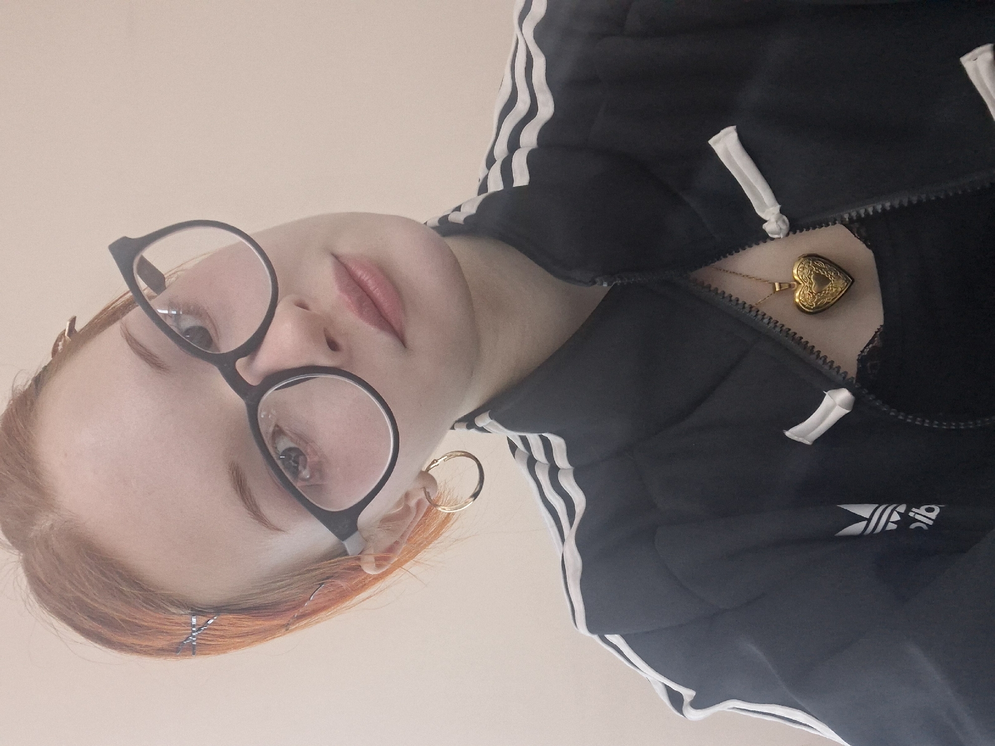 A person with red hair is wearing black-rimmed glasses and a black Adidas track jacket with white stripes. The jacket is partially zipped, revealing a gold heart-shaped locket necklace. There is a small silver hair clip in their hair. They are wearing a gold hoop earring. The image is taken from a low angle looking up. The background is a plain, light color. There is nothing in the photo to indicate the location is Chaikovsky, Russia.