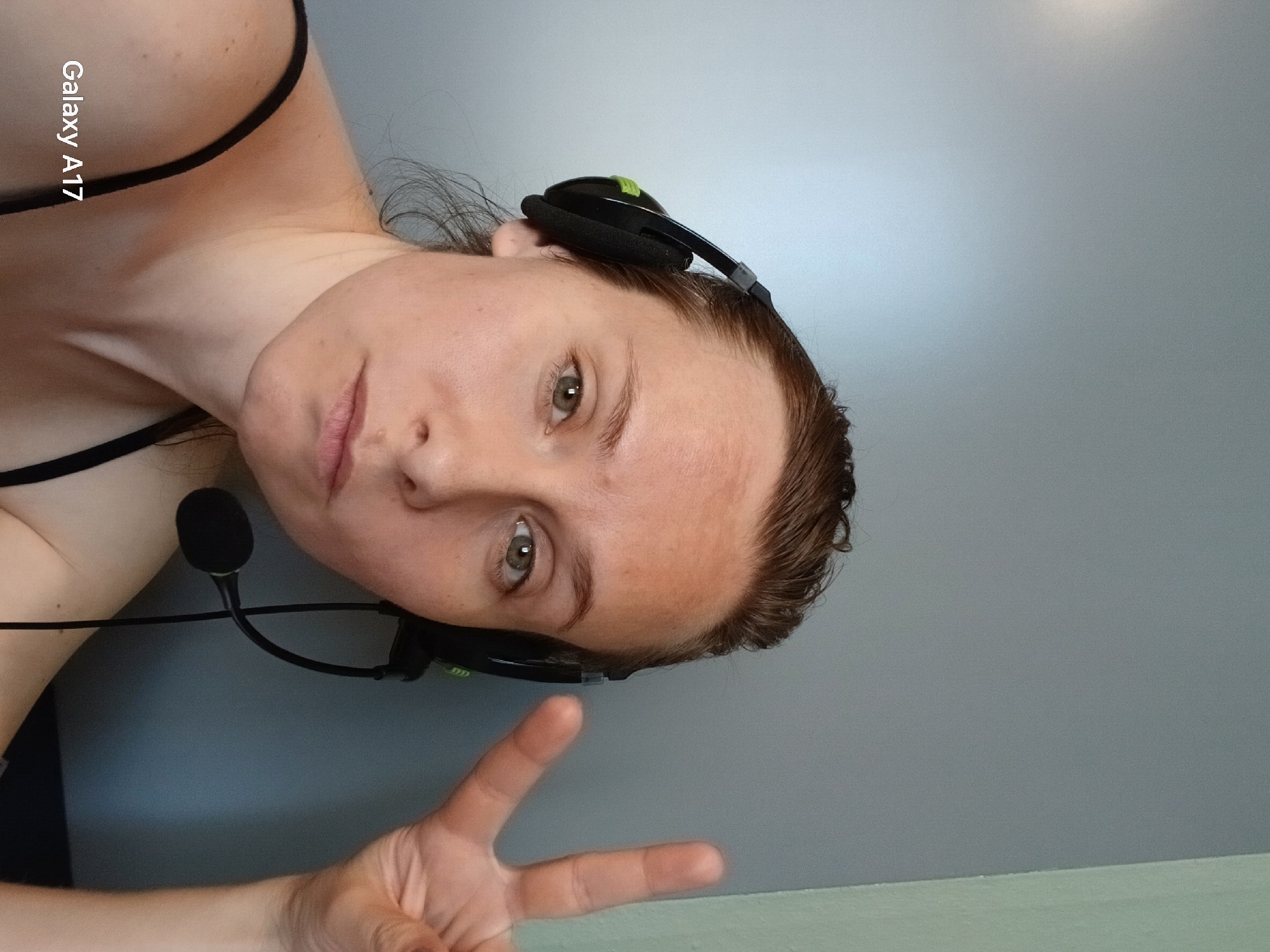 A woman is shown at an extreme angle, possibly self-photographed, wearing a black headset with a microphone. She is making a peace sign with her fingers. The background is a light grey wall. The text "Galaxy A17" is visible in the upper left corner. There is no indication of Yekaterinburg, Russia in the image.
