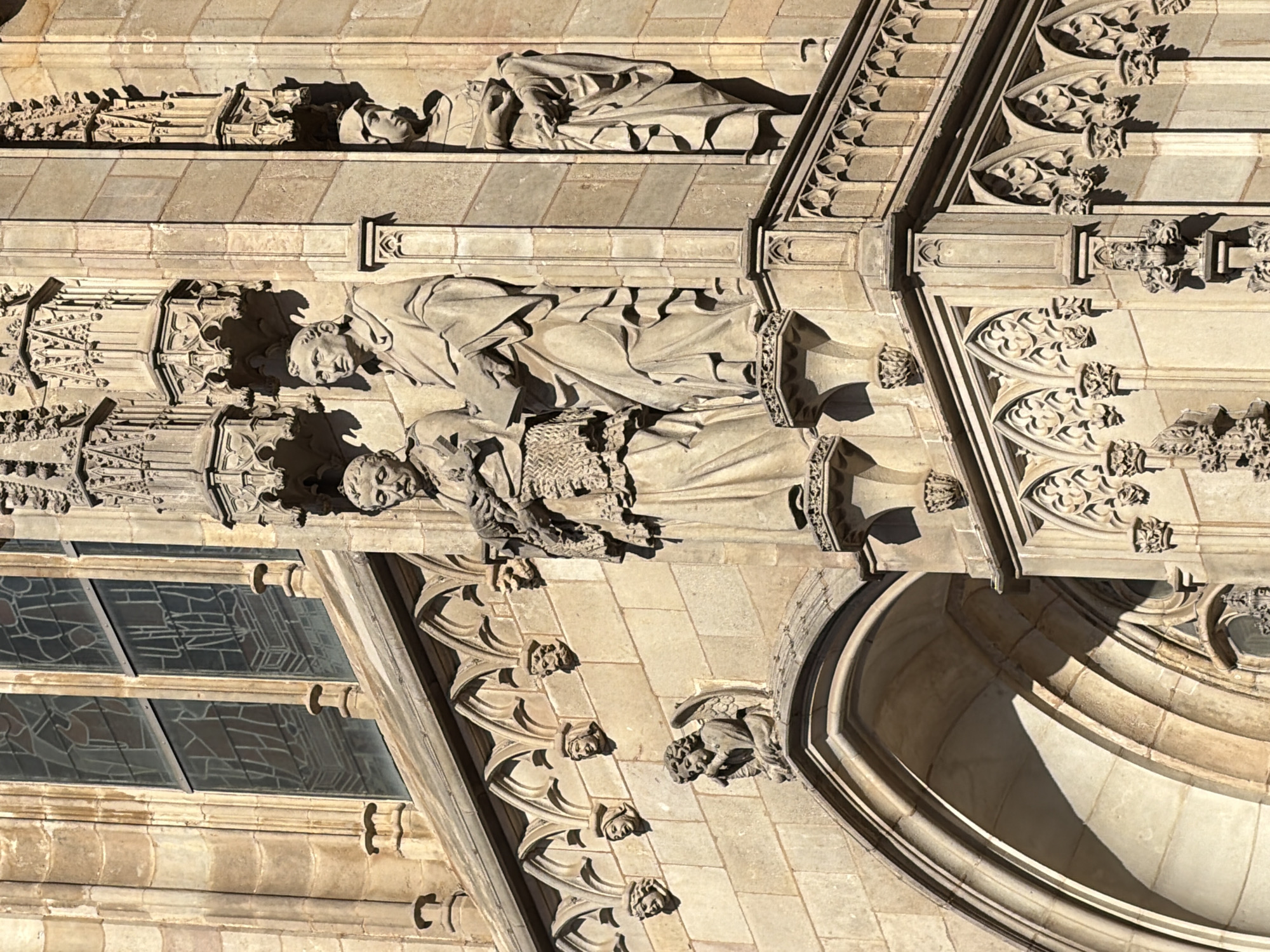 This media file depicts a close-up, vertical view of the facade of a Gothic-style building, likely a cathedral, given the architectural details. The primary subjects are the stone sculptures adorning the exterior. These include two full-figure sculptures of people, one appearing to be a man and the other a woman, both depicted in draped clothing and with serene or solemn expressions. Above these, a reclining figure is visible, also rendered in stone, with drapery creating soft shadows.
The sculptures are integrated into elaborate architectural ornamentation, featuring Gothic tracery, arches, and decorative molding. Small gargoyle-like figures and carved foliage add further detail to the stonework. The building's facade is constructed from light-colored stone, with variations in texture and color suggesting age and weathering.
The scene is illuminated by direct sunlight, casting strong shadows that highlight the three-dimensional quality of the sculptures and the architectural elements. The time of day appears to be midday or early afternoon, based on the intensity and angle of the shadows. The weather is clear, allowing for sharp contrasts and detailed rendering of the stone. The location cues, such as the Gothic architecture, are characteristic of historical European cities, and the prompt specifies Barcelona, Spain. There is no visible text in the image. The overall impression is one of historical grandeur and artistic craftsmanship.