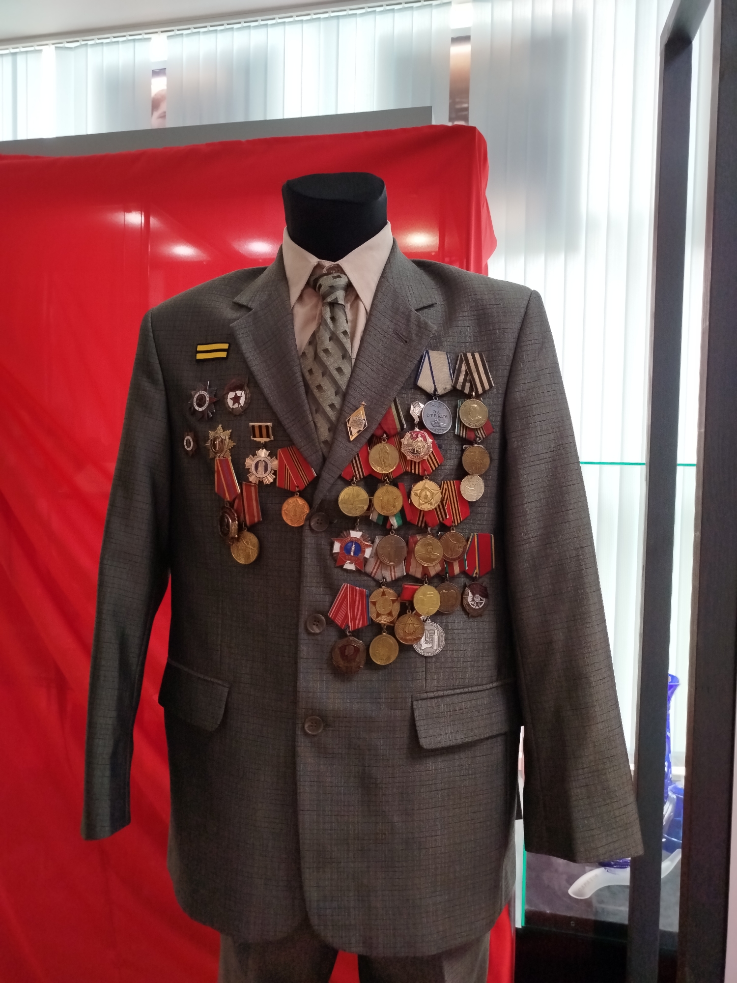 A grey suit jacket adorned with numerous medals and insignia is displayed on a mannequin. The jacket is buttoned, and a patterned tie is visible at the neck. Behind the mannequin, a vibrant red fabric hangs, and to the right, vertical blinds and a dark frame are present. The lighting suggests an indoor setting.