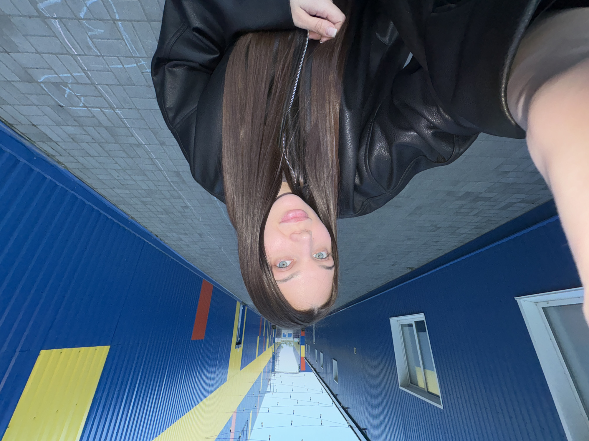 A selfie of a fair-skinned woman with long, dark brown hair and light-colored eyes, wearing a black leather jacket. The background depicts the exterior of an IKEA store, distinguished by its blue corrugated metal walls with yellow and red stripes. Several windows are visible. The location is Chaikovsky, Russia.