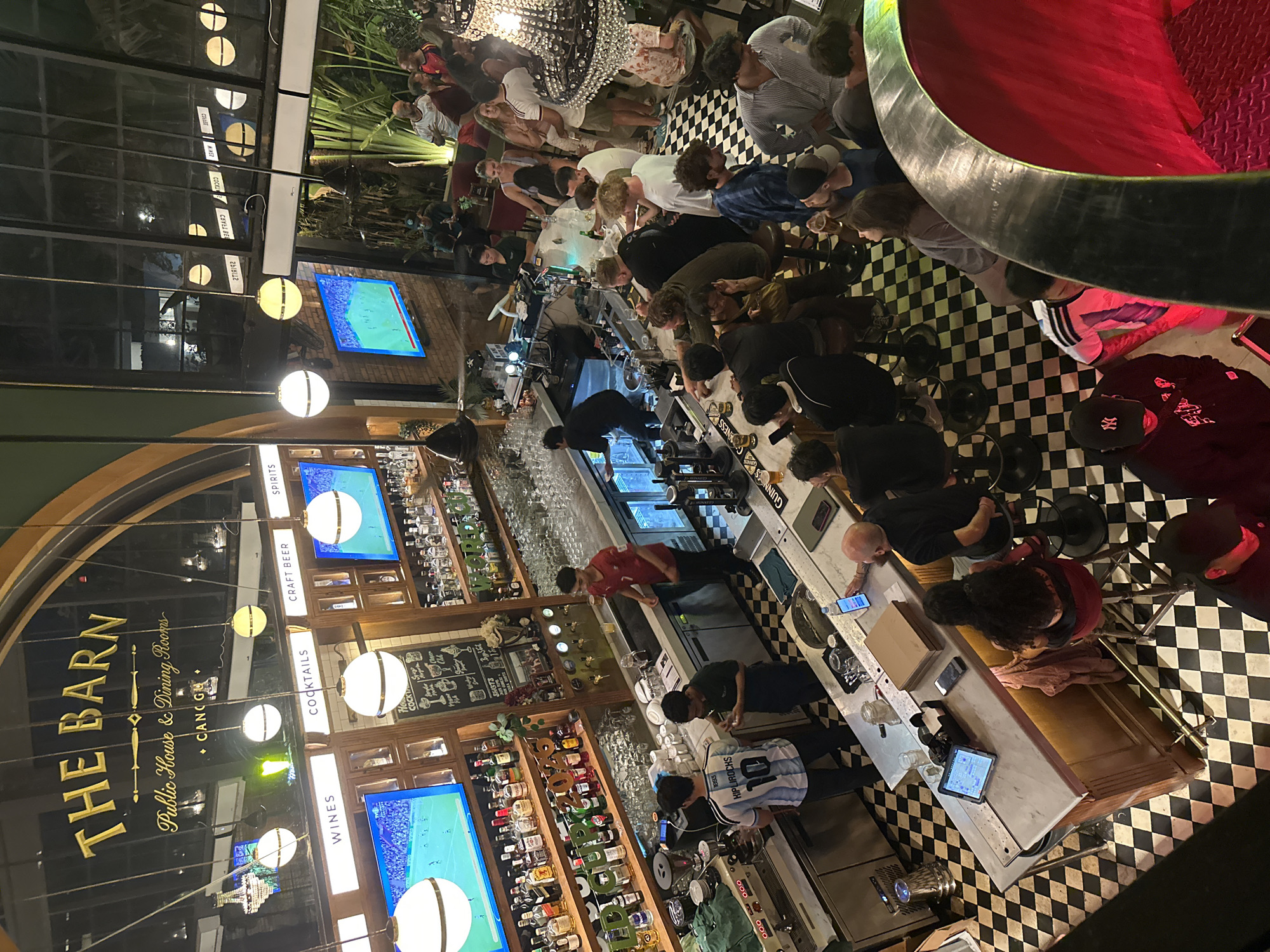 An aerial shot captures a lively scene inside "The Barn," a public house and dining room in North Kuta, Indonesia. The establishment features a long, marble-topped bar with a checkered black and white tiled floor. Behind the bar, shelves are stocked with numerous bottles of alcohol and glassware, with a few television screens displaying sports games. Several bartenders are actively working, preparing drinks and serving customers. Patrons are gathered around the bar, some standing and others seated on stools, engaged in conversation and enjoying beverages. In the background, additional seating areas and decorative elements like chandeliers are visible, suggesting a bustling atmosphere. The overall impression is one of a popular and energetic venue.