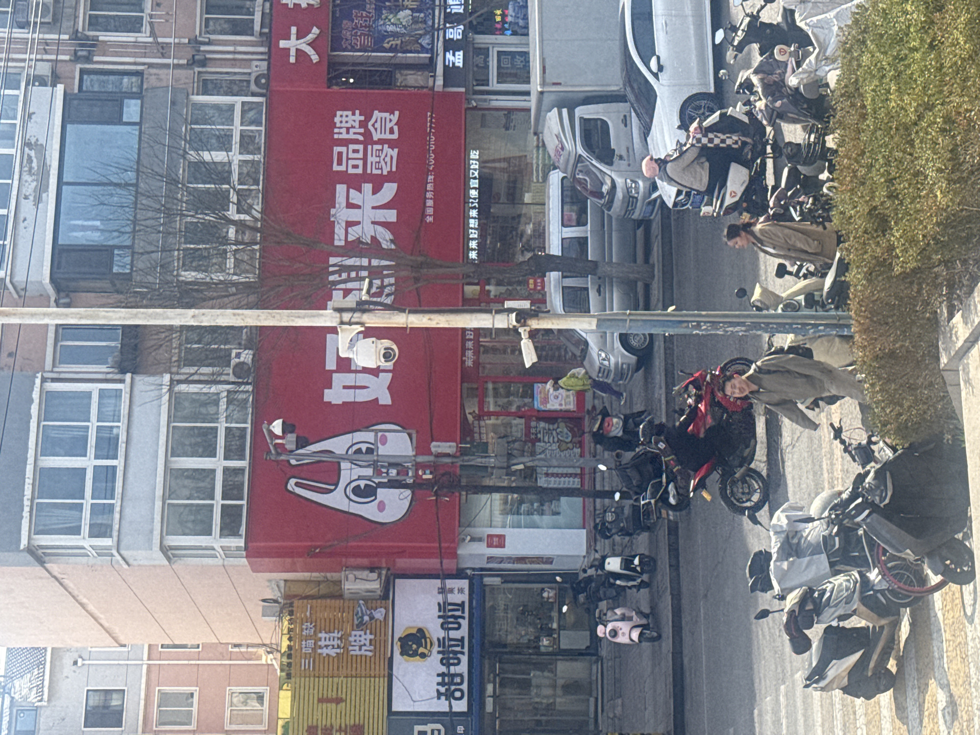 The image shows a street scene in Zhongtun, China. On the left are several multi-story buildings, with signs advertising various businesses. "Hao Lai Lai" is prominently displayed in large red characters. A "Sweet Lola" (or "Tianla La") fresh fruit tea shop can be seen, also with Chinese text. Various motorbikes and scooters are parked along the side of the street. Several pedestrians are visible. Vehicles, including a white van, are also present on the road. Overhead wires run across the street. The environment seems to be urban.