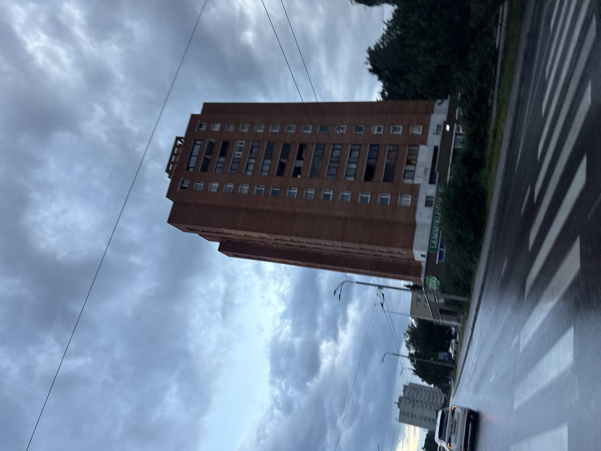 The image depicts a street scene in Tallinn, Estonia, on a cloudy day. A tall, reddish-brown brick apartment building dominates the right side of the frame, with numerous windows and balconies. In the foreground, a wet road with white pedestrian crossing stripes suggests recent rain. A white SUV is visible on the left side of the road, heading away from the viewer. Other buildings, streetlights, and trees are present in the background, indicating an urban environment. A faint sign with the word "LEMMIKKILUPPA" and a green circular logo with "200" is partially visible on the lower part of the brick building. The overall atmosphere is overcast and somewhat somber due to the cloudy sky.