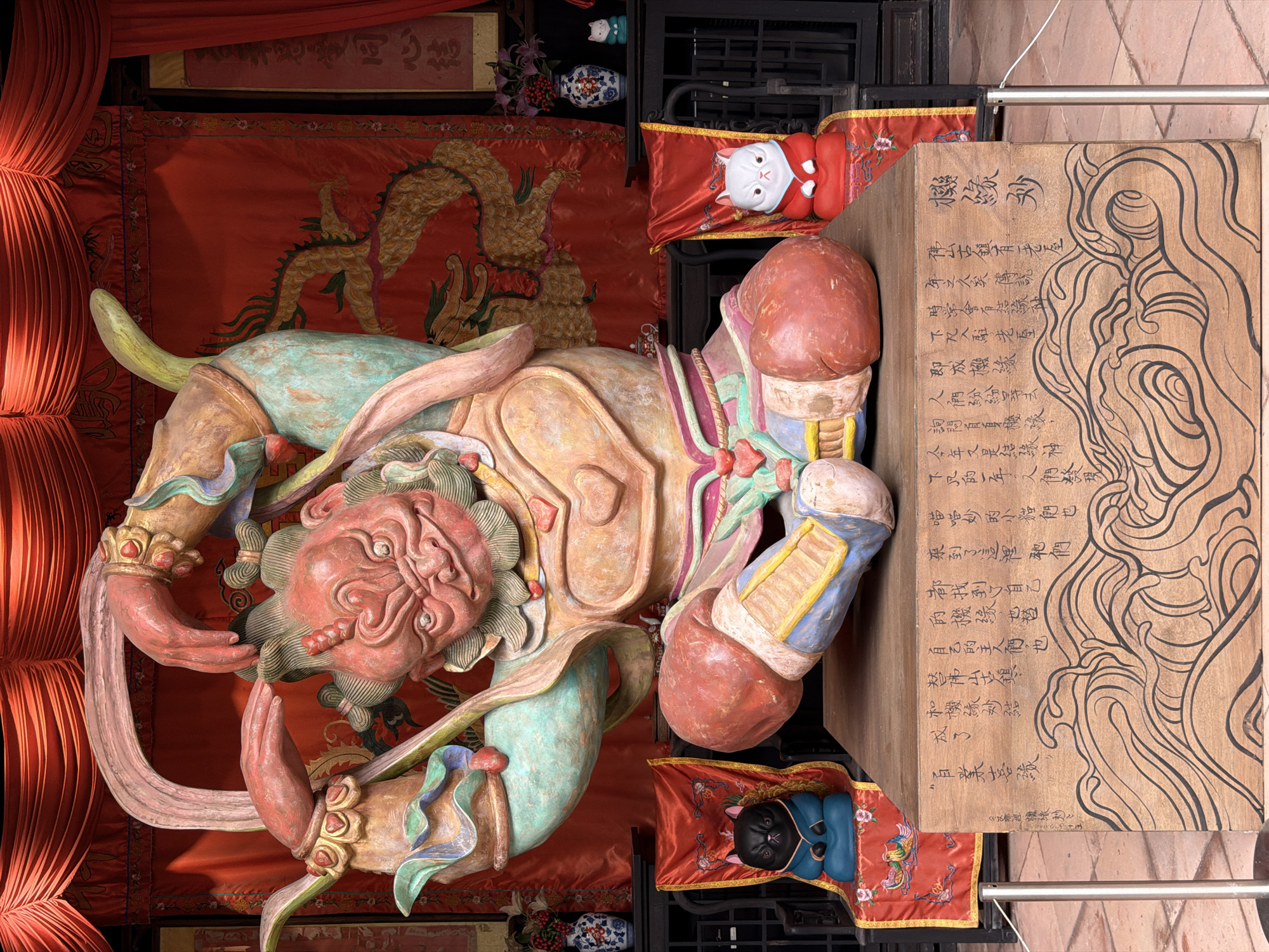An ornate, weathered sculpture of a fierce deity with a red, stern face is the central focus of this image. The deity is adorned in elaborate, colorful armor, with large, muscular limbs posed dramatically. The sculpture rests on a wooden base decorated with carved waves and Chinese calligraphy. Two small, seated cat figurines, one black and one white, are placed on chairs draped with red fabric to the left and right of the sculpture respectively. Behind the deity, a red tapestry with a golden dragon motif hangs, adding to the traditional East Asian aesthetic. The overall setting appears to be an outdoor or semi-outdoor cultural or religious site.
