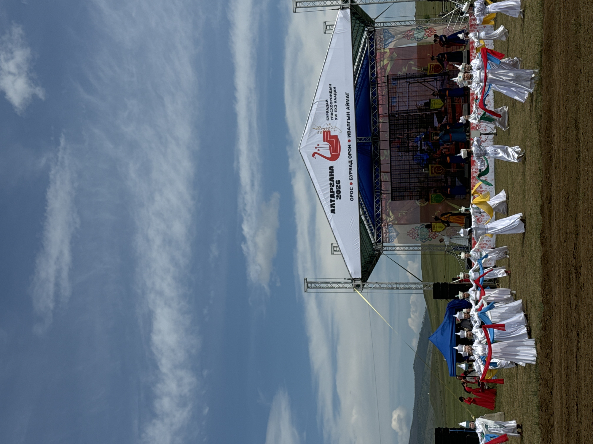 This photograph captures an outdoor cultural performance in Orongoi, Russia. A large, white tent-like structure with signage in Cyrillic script covers a stage. The stage features a screen displaying a colorful, animated scene with traditional figures and banners. On the stage itself, several individuals in traditional dark attire appear to be performing.
In front of the stage, a group of approximately ten people, primarily women, are dressed in white robes and conical hats, holding and maneuvering long, brightly colored sashes of red, blue, green, and yellow. Their movements suggest a choreographed dance. To the left of the main performance area, a smaller group of people in red and white attire are visible, and further left, a blue tent is set up.
The ground is covered in dry grass, with a patch of dark, tilled earth in the foreground. In the background, rolling hills with sparse vegetation and scattered trees extend towards a hazy horizon. The sky is a vibrant blue with scattered white clouds, some wispy and others more substantial. The overall scene is one of a festive outdoor gathering celebrating cultural traditions under a bright, partly cloudy sky.