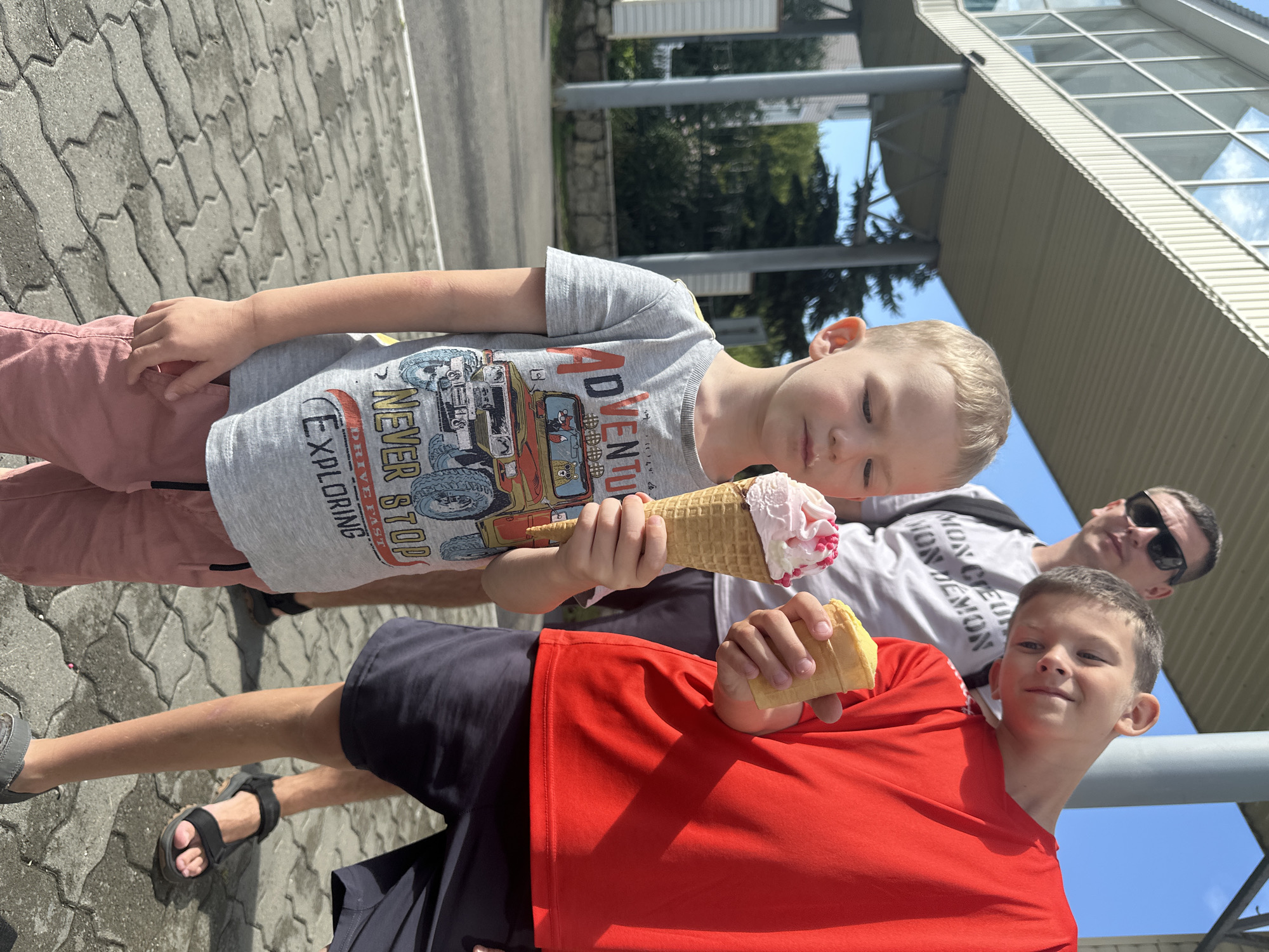 The image captures an outdoor scene with three individuals, two of whom are children, holding ice cream cones. In the foreground, a young boy with blond hair is looking down at his pink and white ice cream cone. He is wearing a gray t-shirt with a graphic of a vehicle and the text "ADVENTURE NEVER STOP EXPLORING". To his right, a slightly older boy is holding a yellow ice cream cone and looking towards the camera with a slight tilt of his head. He is wearing a red t-shirt and dark shorts. Behind them, a man wearing sunglasses and a gray t-shirt with the text "MON CHÂTEAU MON DÉMON" is visible. The background suggests a public area with a paved walkway, a building with a modern architectural design featuring large windows, and some trees and foliage. The lighting indicates a sunny day. The location is identified as Danilovka (Altai Krai), Russia.