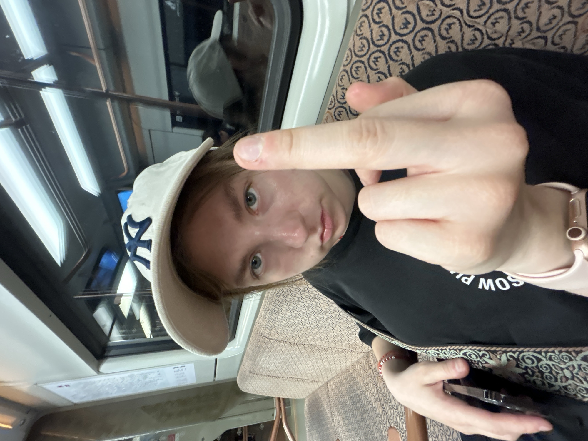 A person is sitting in what appears to be a subway or train car. The person is wearing a white baseball cap with a blue logo. The person is also wearing a black shirt with white lettering. The person is holding up their middle finger. A watch is visible on the person's wrist. The subway car has a window and patterned upholstery. It is unclear if the person is in the city of Unknown, Unknown.