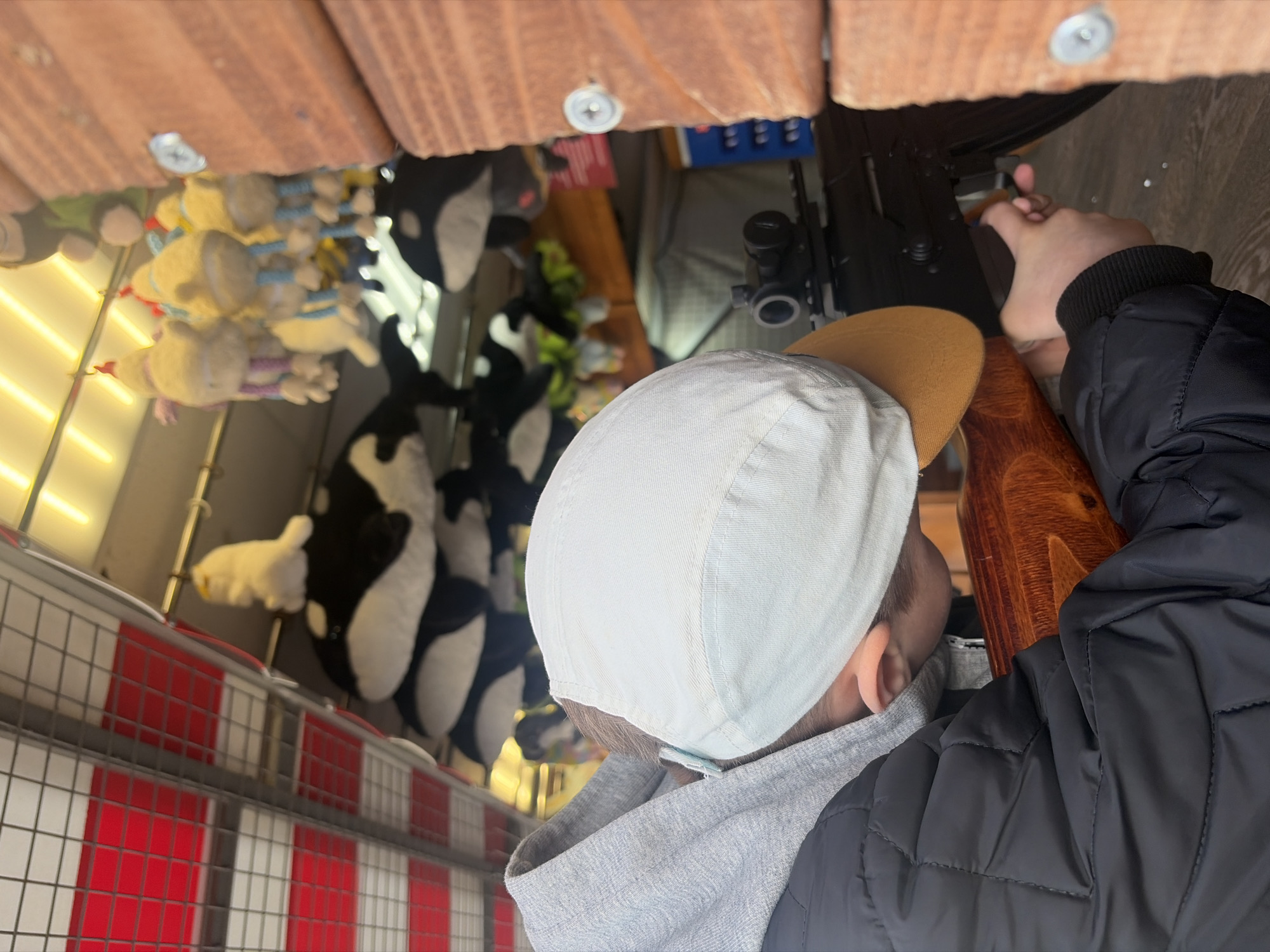 A person in a light-colored baseball cap and black jacket is shown operating a rifle at a carnival shooting range. The range is lined with stuffed animals as prizes, including orca whales and yellow characters. The background includes a red and white barrier fence and overhead wooden beams. The location cannot be discerned to be Saint Petersburg, Russia.
