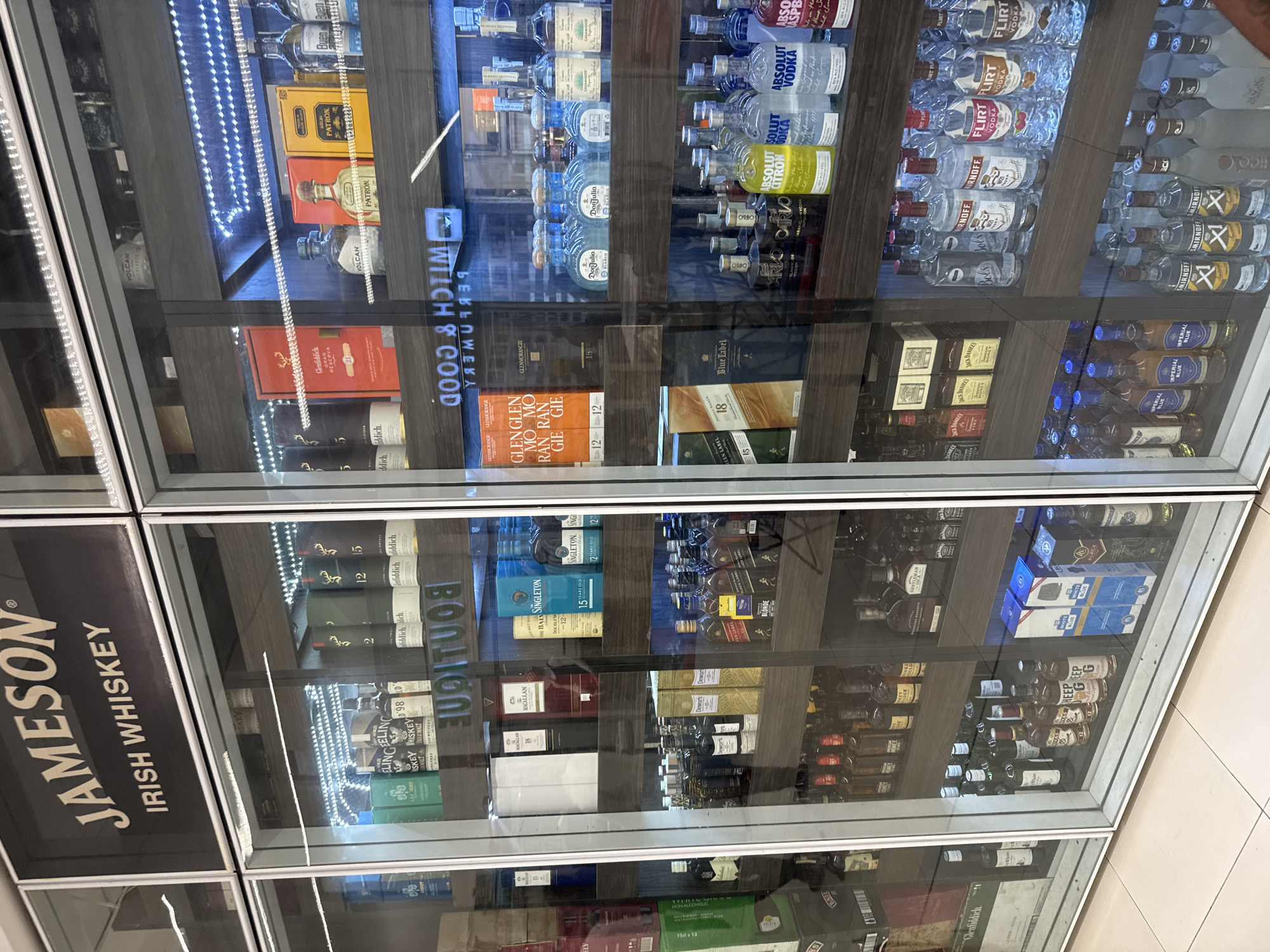 The image shows a display case filled with various alcoholic beverages, including whiskey, vodka, tequila, and other liquors. Prominent brands visible include Jameson Irish Whiskey, Glenfiddich, Macallan, Glenmorangie, Patron, Don Julio, Absolut Vodka, Smirnoff, Flirt Vodka, Jack Daniel's, and Johnnie Walker Blue Label. The sign "WITCH & GOOD" is reflected on the glass. The display case appears to be located in a retail environment, likely a liquor store or supermarket in Abuja, Nigeria.