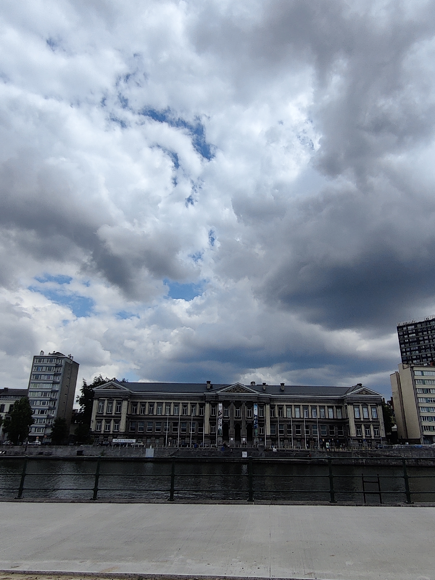 The image depicts the Quai Malaise in Liège, Belgium, under a dramatic, cloudy sky. The focal point is a grand, neoclassical building with a long facade featuring numerous windows and ornate architectural details. This building appears to be a cultural institution, possibly a museum or university building, as suggested by signs and figures visible on the facade. In the foreground, a dark metal railing runs along the edge of a wide river, likely the Meuse. Across the river, a modern, high-rise apartment building stands out against the sky, contrasting with the historical architecture. The scene is set on a paved promenade or walkway, suggesting a public area along the riverfront. The overall impression is one of urban life with a blend of historical and contemporary elements under a moody sky.