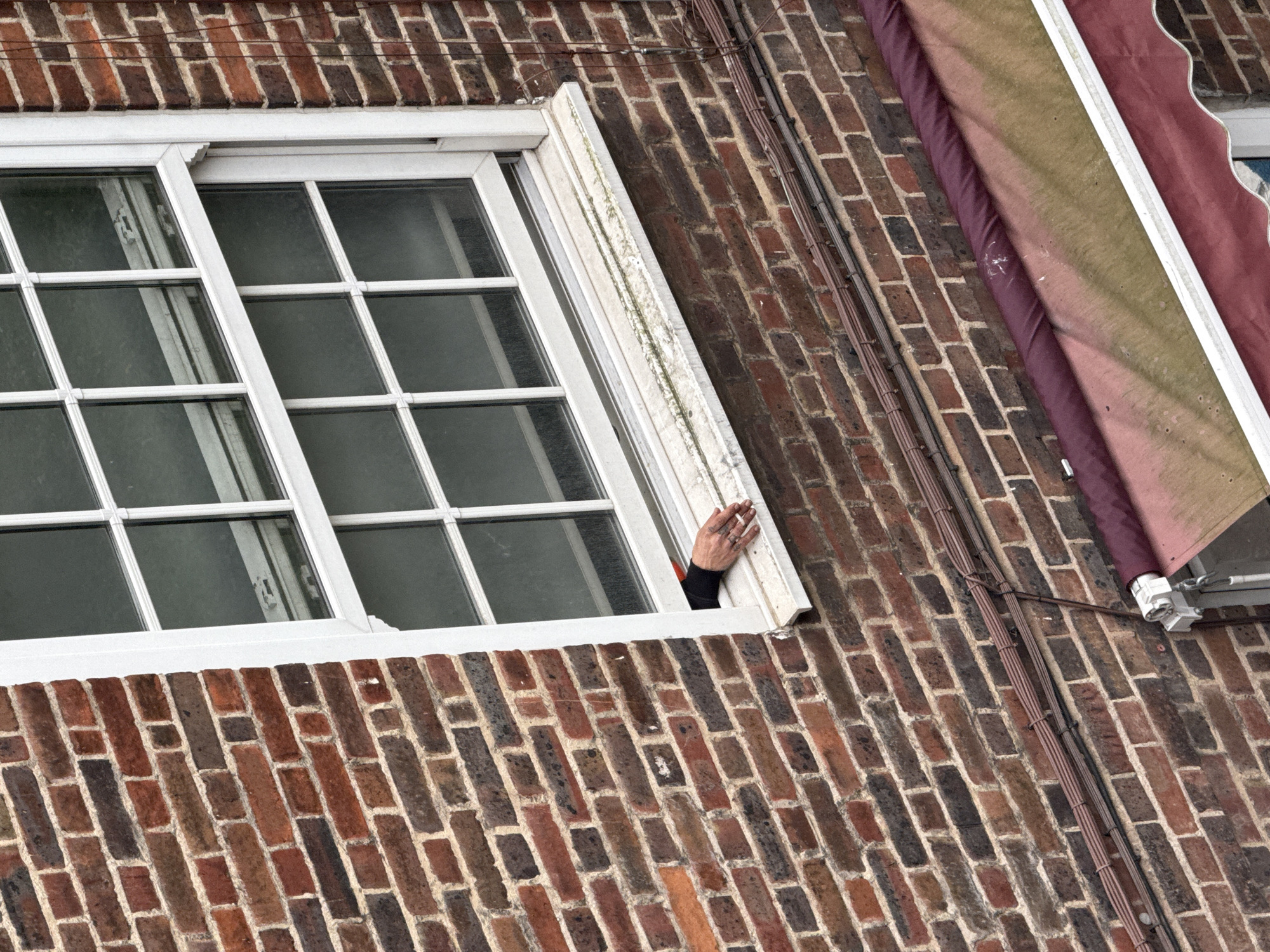 A medium shot, taken from a slightly low angle, shows a window in a brick building. The window, which is open, is white with a multi-pane design. A hand extends out of the window, holding a cigarette. The arm is covered in a black sleeve. The brick building features reddish-brown bricks with darker bricks interspersed. A brown and burgundy awning is visible to the right of the window, with electrical cables running down the brick wall. The overall composition suggests an urban setting.