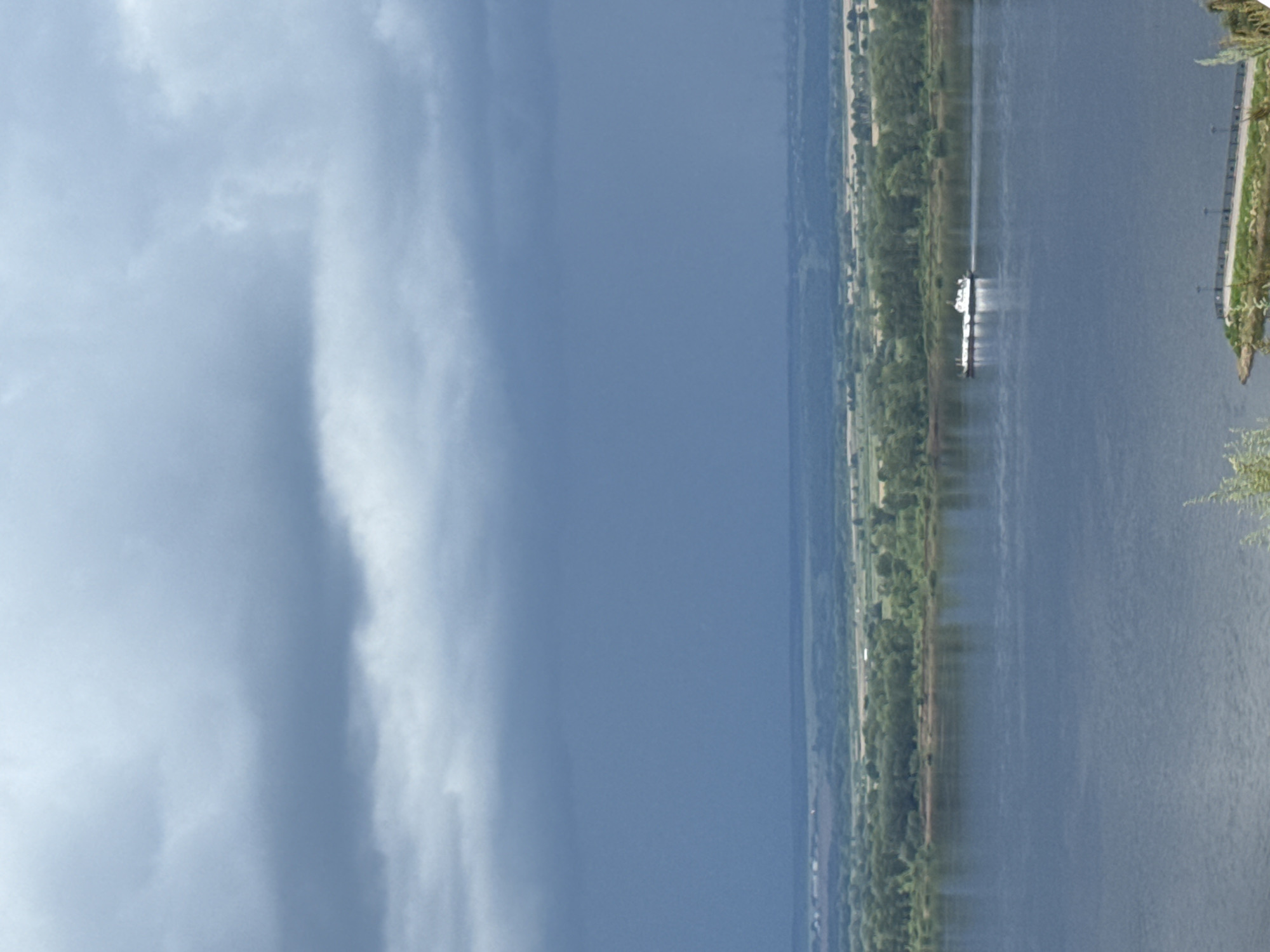 The image depicts a wide river with a barge moving from left to right in the middle distance. The barge is white with a long, dark cargo. The river is calm, reflecting the overcast sky. On the far bank of the river, there is a strip of green trees and then a hazy, rolling landscape stretching to the horizon. The sky is filled with dark, dramatic clouds, suggesting an impending storm. In the foreground, a small portion of a rocky embankment with a walkway and lampposts is visible, with some greenery and foliage in the lower right corner. The scene is consistent with the views from the upper bank of the Kama River in Yelabuga, Russia, which is known for its dramatic river vistas and cloudy skies. There are no people visible in the image.