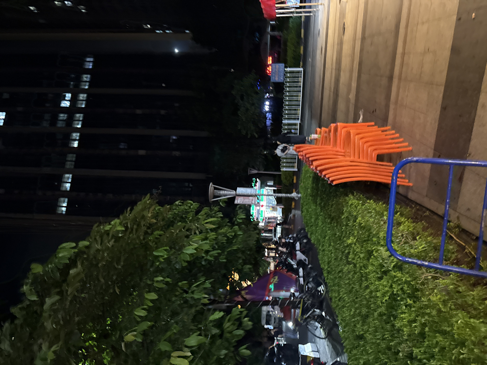 In the Haizhu District of China, at night, a street scene unfolds. A dense, green hedge lines the foreground, with a blue metal barrier partially visible on the left. To the right of the hedge, a row of bright orange plastic chairs sits empty, facing the viewer. Beyond the chairs, a paved walkway extends, illuminated by streetlights. In the midground, several parked motorcycles and bicycles are visible, some with purple flags attached. A large tree with lush green foliage dominates the left side of the image, its leaves reflecting ambient light. Behind the trees and motorcycles, a tall, dark building with numerous illuminated windows rises into the night sky. A street lamp stands near the hedge, and a few people are scattered throughout the scene, mostly in the midground, appearing to be walking or standing. The overall impression is of a quiet, urban setting at night, with a mix of natural elements and man-made structures.