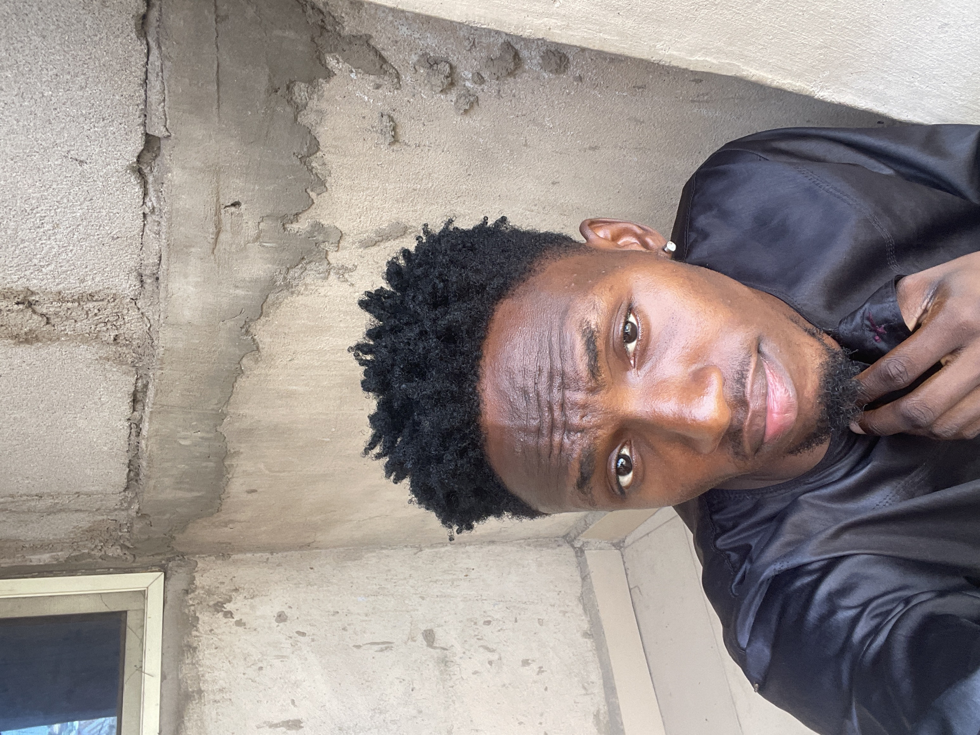 The image is a portrait of a young man, appearing to be a selfie, captured in daylight. He has dark skin, a short, dense crop of black curly hair, and a neatly trimmed beard and mustache. He looks directly into the camera with a focused, slightly serious or contemplative expression, with a subtle furrow in his brow. He wears a black, long-sleeved garment that has a subtle sheen, suggesting it might be made of satin or a similar fabric, possibly a traditional tunic. His right hand is partially visible near his chest.
The background primarily consists of unpainted concrete walls displaying varied textures, some areas appear rough and worn with visible cracks and patches, suggesting an older or unfinished structure. In the lower-left corner of the frame, a section of a simple white-framed window with a mesh screen is visible. The bright, diffused lighting indicates that the photo was taken during the day under clear conditions. The setting appears to be a humble, functional outdoor or semi-outdoor space. There is no visible text in the image. The architectural style, particularly the concrete walls, is consistent with common building types in regions like Lokoja, Nigeria.
