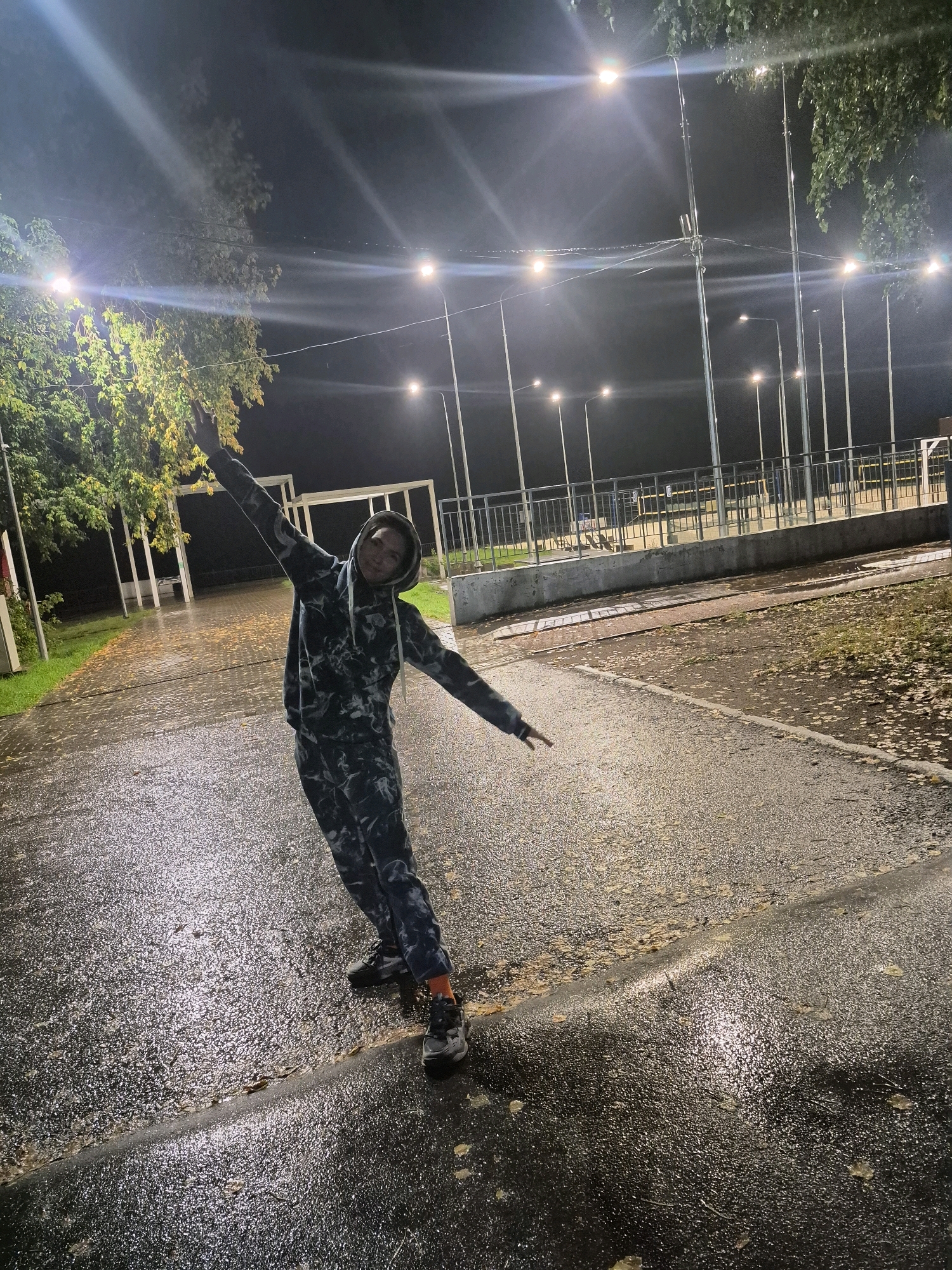 A person stands on a wet, paved surface at night, illuminated by streetlights. The person is wearing a dark camouflage-patterned hooded tracksuit and grey sneakers with orange socks visible. They are in a dynamic pose with arms outstretched, leaning to their right. The ground is reflective and appears wet from rain, with fallen leaves scattered on it. In the background, there are trees with some leaves, lampposts, a metal fence, and structures that resemble bleachers or shelters. The overall scene suggests an outdoor recreational area or sports field at night in Chaikovsky, Russia, possibly after rainfall.