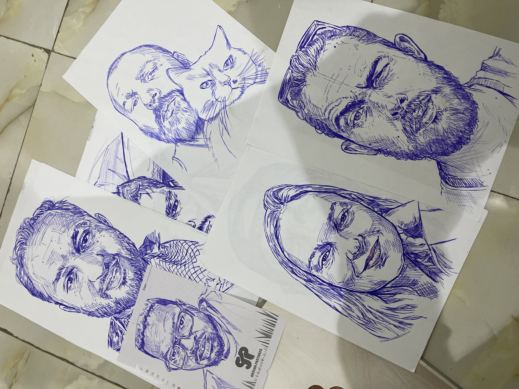 The image shows a top-down view of several ballpoint pen sketches of people and a cat, laid out on a tiled floor. The sketches are drawn on white paper. In the upper portion of the image, a sketch of a man with a beard and a collared shirt is visible. Below and to the left of this, is a smaller sketch of a man wearing glasses, with a logo that reads "SP GREENA PASTURES". To the right of the first man's sketch, a sketch of another man with a beard is seen, with a cat resting its head on his shoulder. In the lower portion of the image, a sketch of a woman with long hair and a smiling expression occupies the left side, while a sketch of a man with a beard and sunglasses perched on his head is on the right. The floor appears to be made of light-colored, polished tiles with a subtle marble-like pattern. There are no visible indications of the city of Abuja, Nigeria, or any specific environmental details that would pinpoint the location.