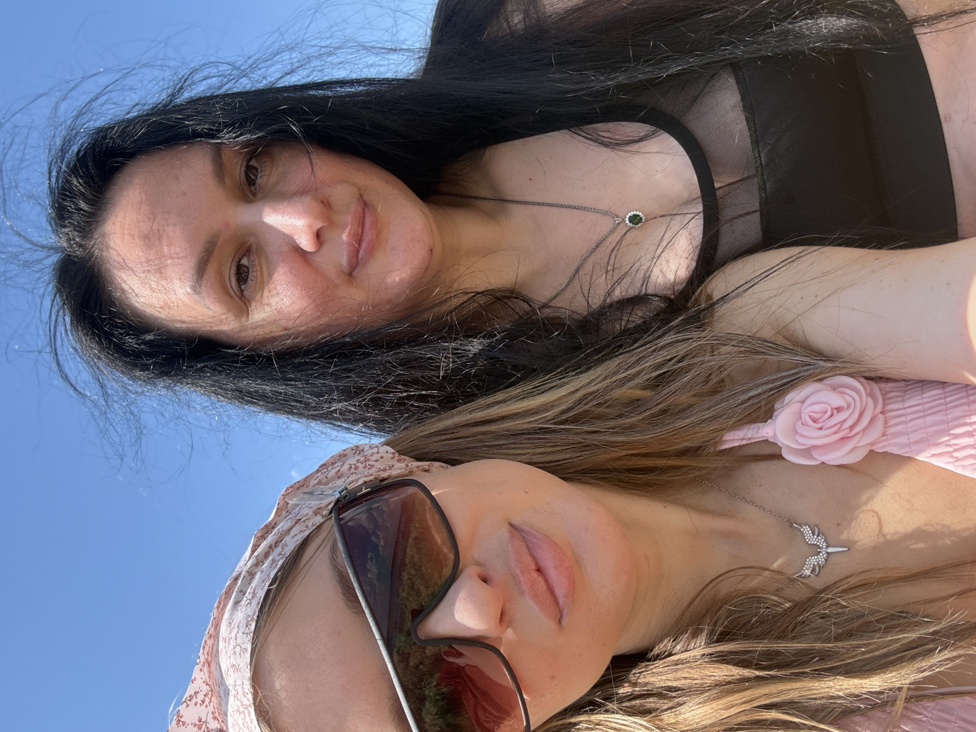 Two women are captured in a selfie taken outdoors under a clear blue sky. The woman on the left, wearing a pink top and a floral bandana, has blonde hair and is sporting large sunglasses. She is adorned with a delicate necklace featuring a winged pendant. To her right, a woman with long, dark hair is wearing a black mesh top and a silver necklace with a green pendant. The background suggests a rural or natural setting.