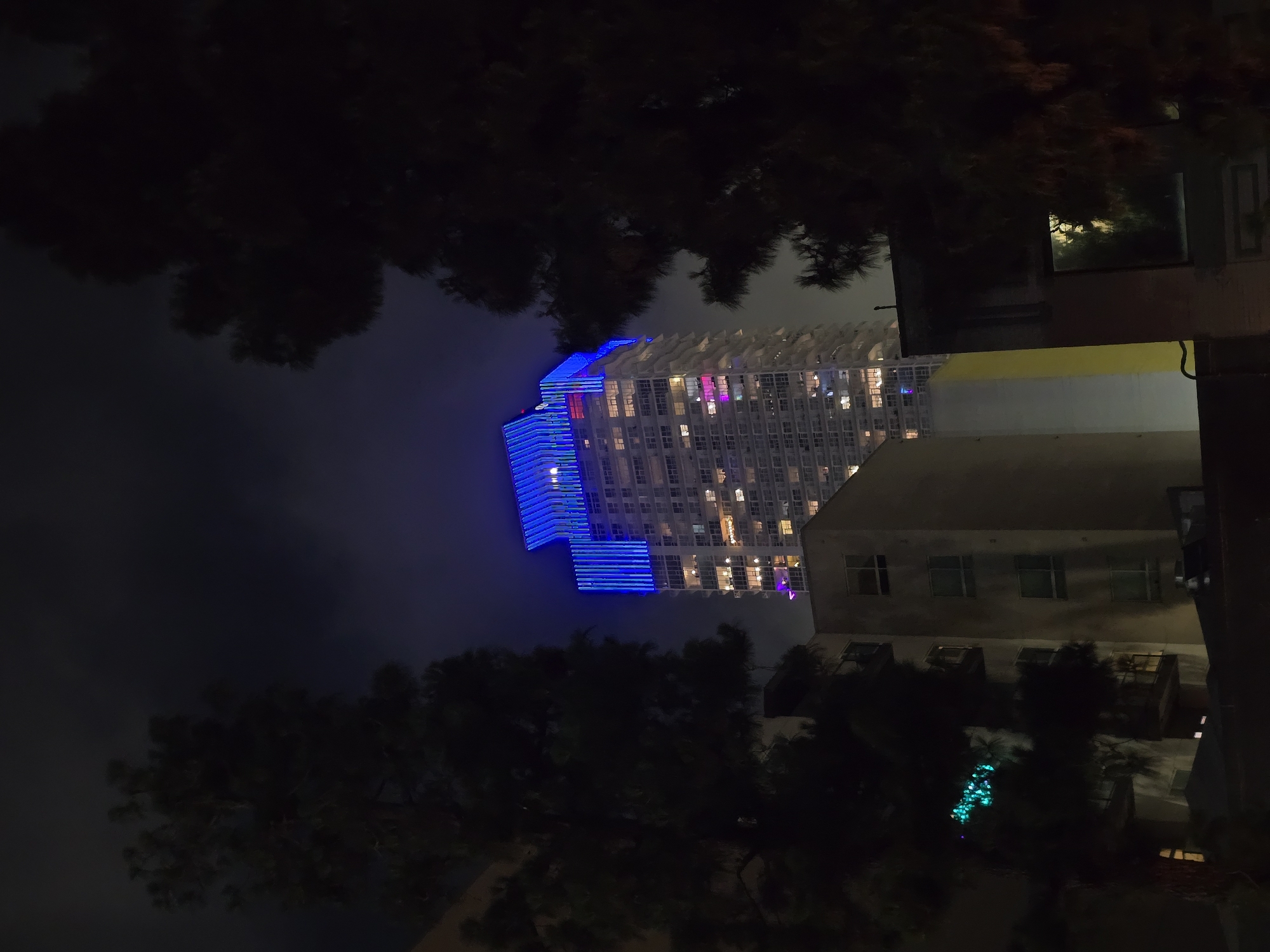 An outdoor, nighttime, eye-level, medium shot captures a modern, multi-story building illuminated with blue lights on its upper floors, situated in an urban setting. The building is partially obscured by the dark silhouettes of trees in the foreground. The building's windows are lit, suggesting occupancy. Other structures, including a lower, beige building with visible windows and balconies, are visible to the left and center of the frame. The sky is dark, with hints of clouds. The overall ambiance is one of city nightlife.