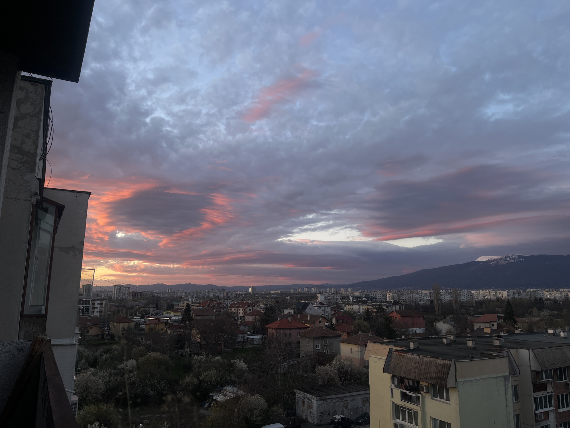 The image shows an elevated view of Sofia, Bulgaria, during a colorful sunset or sunrise. The sky is filled with clouds streaked with pink and purple hues, indicating either the beginning or end of the day. The landscape is dominated by a dense urban environment consisting of residential buildings, houses with red and grey roofs, and scattered trees. In the distance, a mountain range with patches of snow is visible. The photograph is taken from a high-rise building, as the side of the building can be seen on the left side of the image. The location is somewhere in Sofia, Bulgaria, judging by the architecture and the presence of Vitosha Mountain in the background. There are no people visible in the image. The atmosphere is still and calm.
