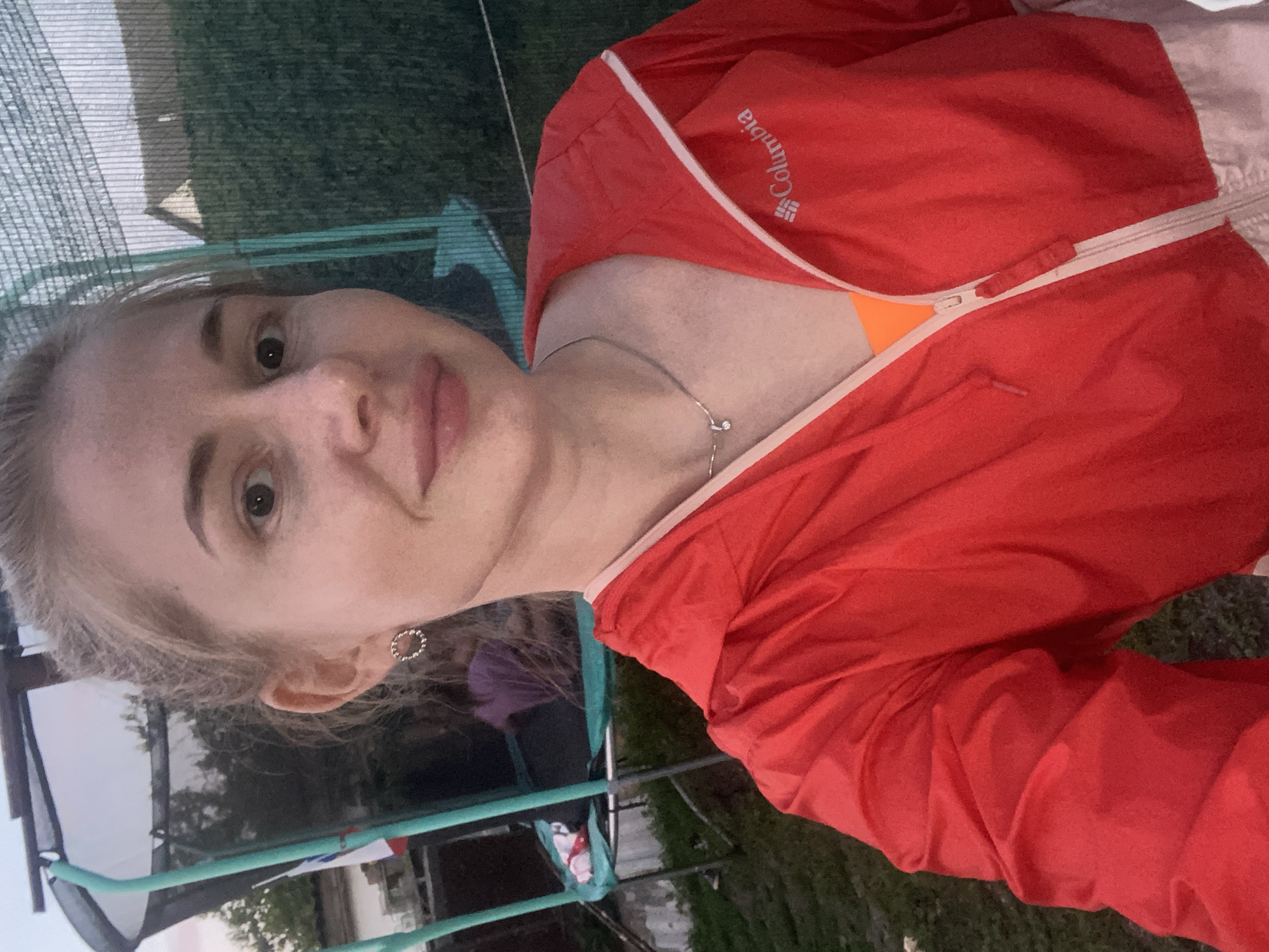 The media file shows a young woman taking a selfie outdoors. She is wearing a red hooded jacket with a white zipper and a light-colored lining. The jacket has the Columbia logo visible on the upper left side. She is also wearing an orange top underneath. The woman has blonde hair pulled back and is wearing a small, circular earring on her left ear.
The setting appears to be a grassy area, possibly a backyard, with what looks like a trampoline frame and netting in the background. Trees and some structures are visible in the distance, suggesting a suburban or rural environment. The lighting indicates it is either early morning or late afternoon, with soft, diffused light. The woman's expression is neutral, and she seems to be casually posing for the photo. There are no other people prominently featured, though a blurry figure in a purple shirt can be seen in the background.
The orientation of the image is portrait mode, with the woman's face taking up a significant portion of the frame. The overall mood is relaxed and informal. The location context provided is Маринопіль, Ukraine.