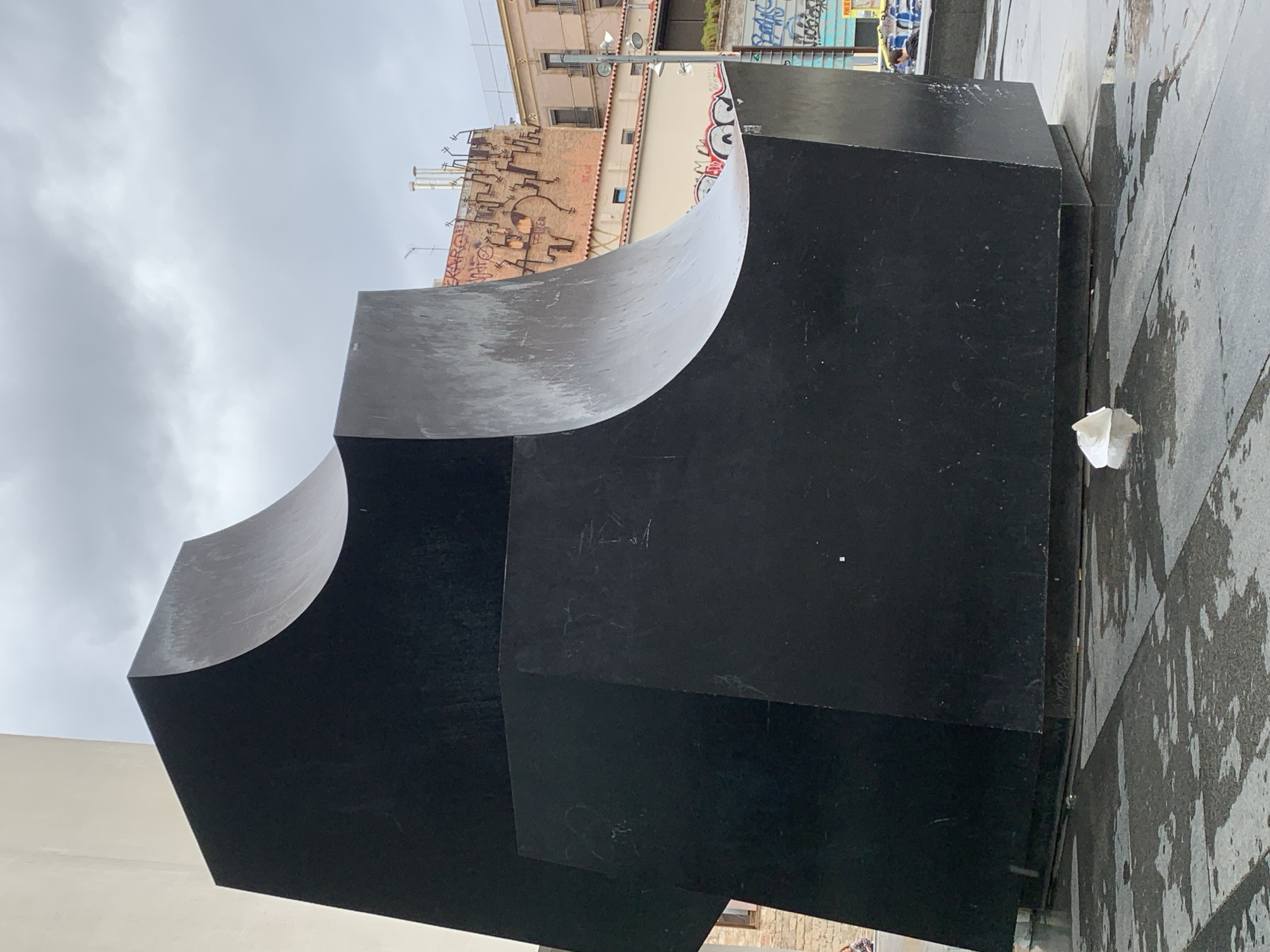 The image shows a large, abstract, matte-black sculpture composed of cubic and cylindrical elements. The sculpture is situated on a paved surface, likely a pedestrian area, with wet stone tiles visible. Behind the sculpture, a building facade is visible, displaying a mix of architectural styles and graffiti. The sky is overcast, suggesting a cloudy or rainy day. A small piece of crumpled white paper lies on the ground near the sculpture. The location is Barcelona, Spain, confirmed by the architectural style of the buildings in the background. No people are directly visible in the immediate vicinity of the sculpture, but hints of people can be seen near the top right.