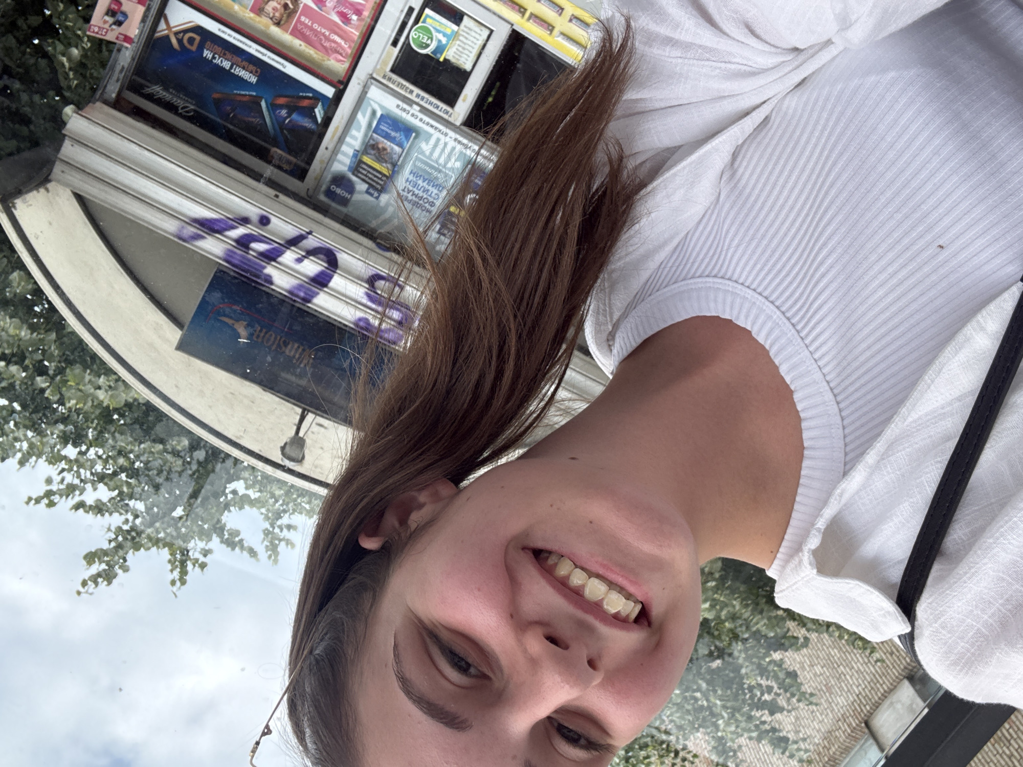 The image is a selfie taken by a young woman with long brown hair, wearing a white ribbed top and a white open shirt, with a black strap visible across her chest. She is smiling, showing her teeth. Behind her, through what appears to be a window, is a kiosk displaying various tobacco products, with text visible in Bulgarian. The kiosk is situated on a street in Burgas, Bulgaria. Trees and a cloudy sky are visible in the background.