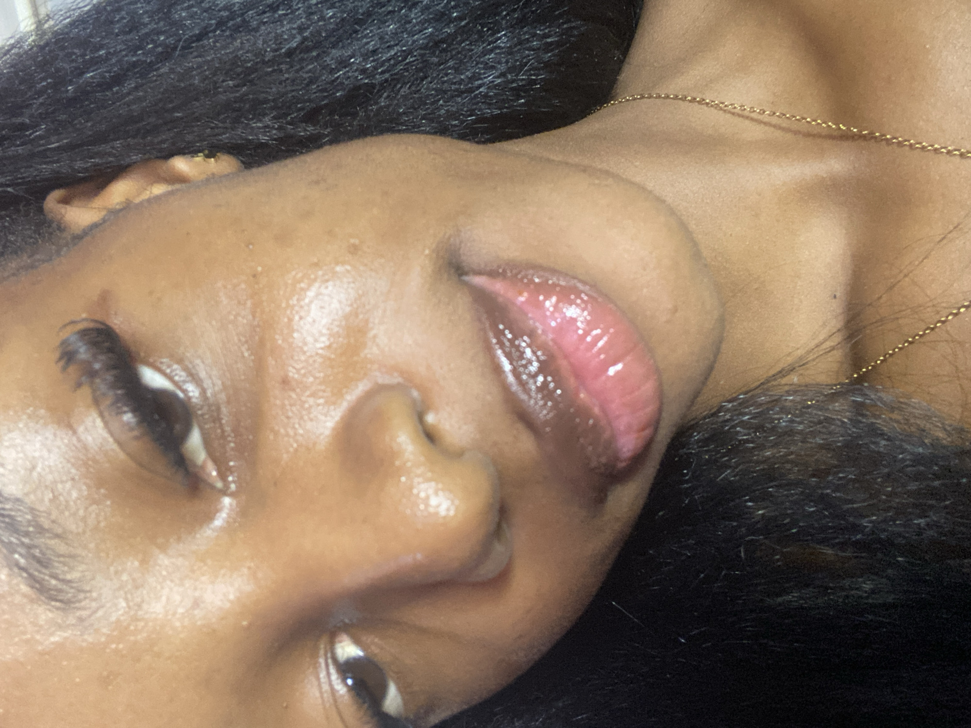 The image shows a close-up, low-angle, eye-level, indoor shot of a young Black woman's face, with her right ear and neck visible. She has dark hair, dark eyebrows, and is wearing mascara and lip gloss. Her skin appears to be glowing, possibly due to sweat or a dewy makeup product. A gold chain necklace is visible around her neck. The background is dark and out of focus, suggesting an indoor setting. There is no indication of the city of Jos, Nigeria, or any specific location within it.