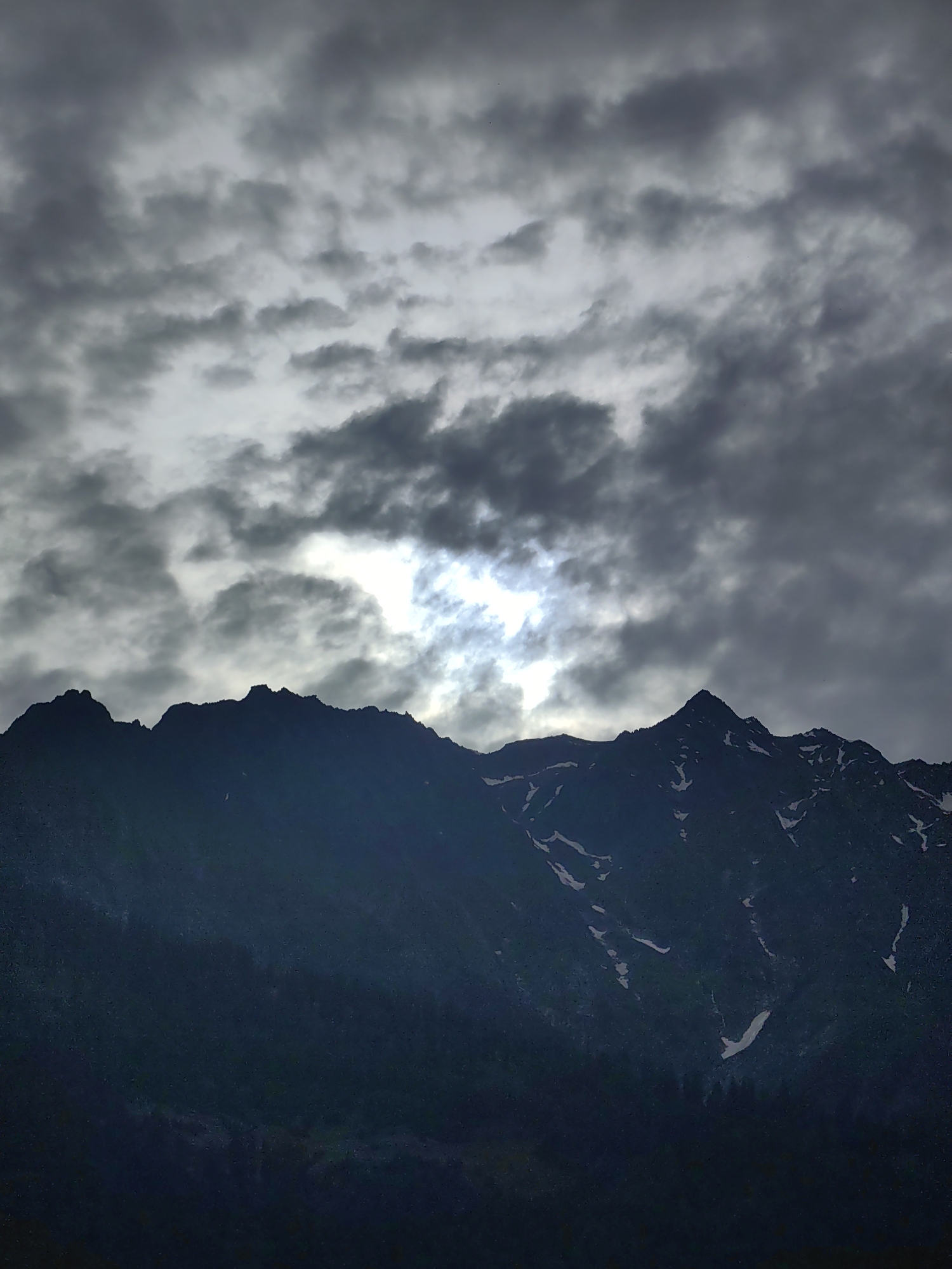 The image depicts a dramatic mountain landscape under a heavily clouded sky. The foreground is dominated by the dark silhouette of a rugged mountain range, with some patches of snow visible on its upper slopes. The texture of the snow suggests a cold, high-altitude environment. The clouds above are dark and ominous, with the sun's rays breaking through in a bright, diffused area, creating a strong contrast. The overall mood is somber and majestic. This scene is likely located in or near Vashisht, India, a popular tourist destination known for its mountainous terrain and spiritual sites.