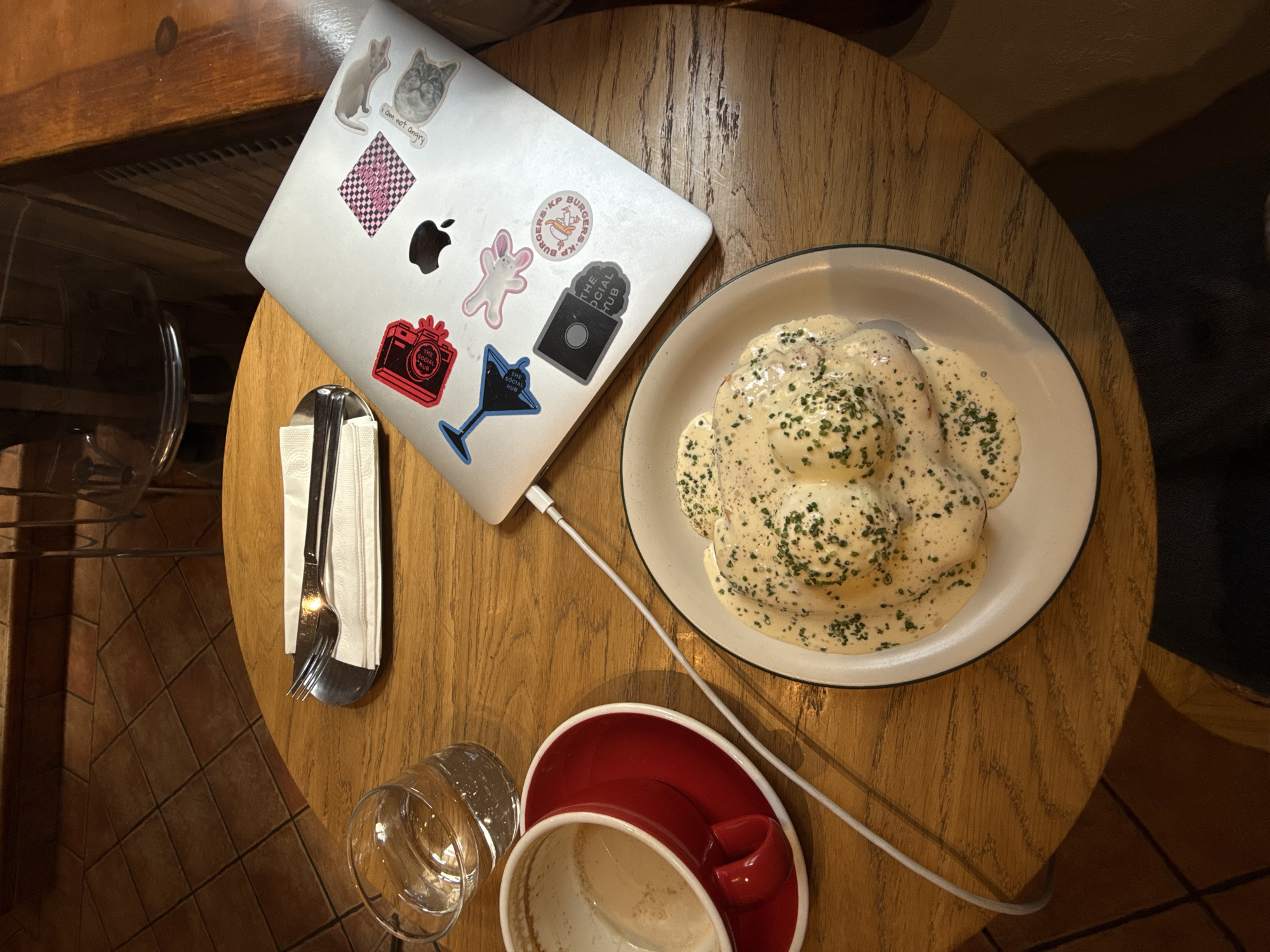 An overhead shot captures a cozy scene on a round, wooden table, likely in a cafe or restaurant in Belgrade, Serbia. The warm, dim lighting suggests a relaxed atmosphere.
A silver laptop is prominently placed on the left side of the table, open and connected by a white charging cable. The laptop lid is adorned with several stickers: a light brown cat and a grey cat sticker with the text "i am not angry," a black and pink checkerboard pattern, a black Apple logo, a pink bunny-like figure, a red camera with "THE SOCIAL HUB" written on it, a blue martini glass also bearing "THE SOCIAL HUB," a black rectangular sticker with "THE SOCIAL HUB," and a partially visible circular sticker that reads "BURGERS KP B BURGERS KPBU."
To the right of the laptop, a white plate with a dark rim holds a dish covered in a rich, creamy white sauce generously sprinkled with green herbs, possibly chives. The food beneath the sauce appears to be a collection of rounded, soft-textured items.
Towards the bottom left of the table, a clear glass is visible, and next to the laptop, a fork and spoon are neatly wrapped in a white napkin. An empty red coffee cup with a matching saucer, showing residual coffee stains, sits at the bottom center of the frame. The floor beneath the table, visible in the bottom left, is covered with reddish-brown square tiles.
The arrangement suggests an individual is engaged in work or leisure on their laptop while enjoying a meal and a beverage, likely coffee, in a comfortable setting.