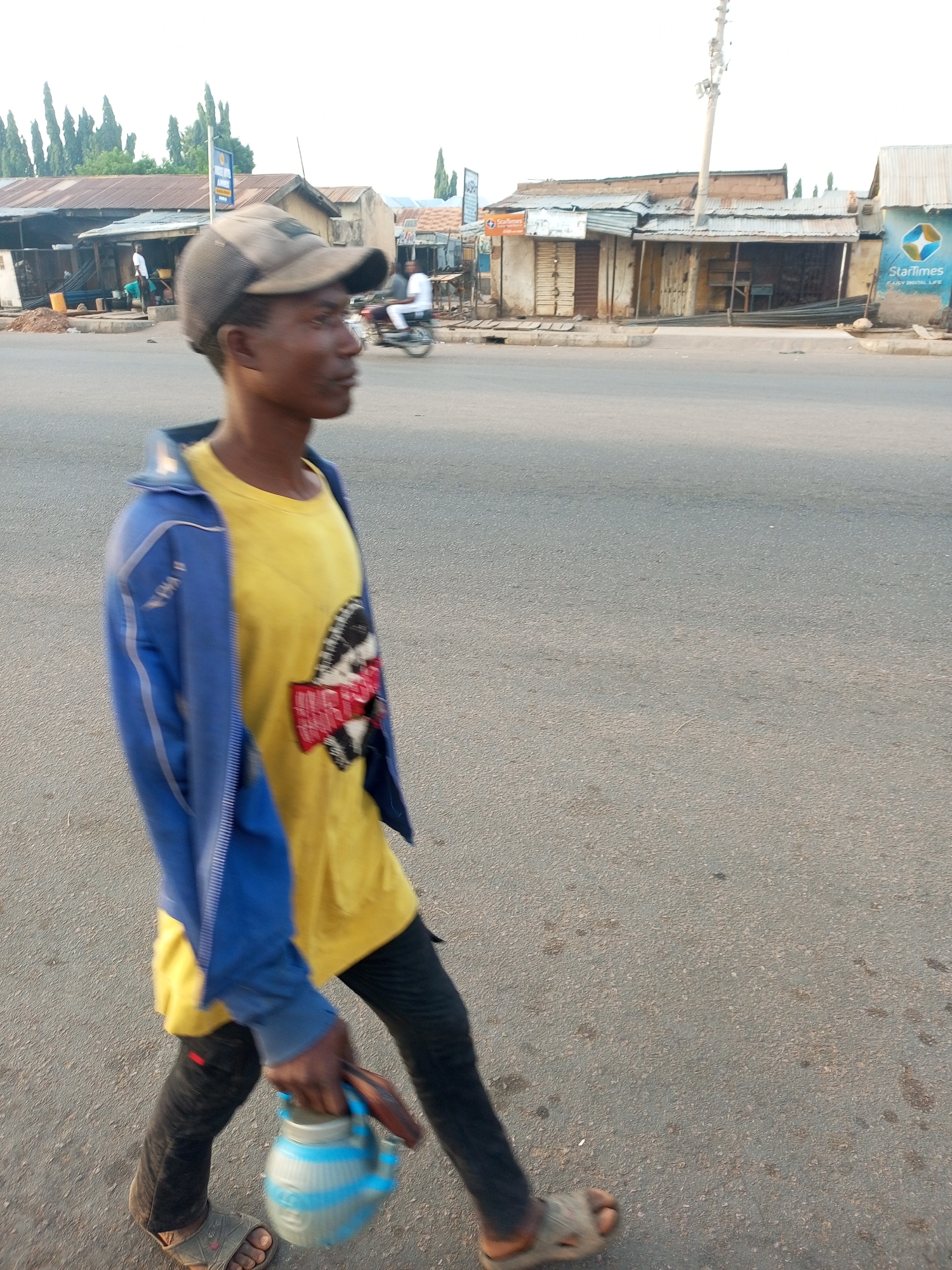 The image shows a dark-skinned man walking on a paved street. He is wearing a grey baseball cap, a yellow t-shirt with a design, a blue jacket, black pants, and sandals. He is carrying a blue and white container with a handle in his left hand. The background features a street with buildings on either side. The buildings appear to be shops or businesses with corrugated metal roofs. Signs and logos are visible on some of the buildings, including "StarTimes." Other people and a motorbike can be seen in the background. The image appears to be taken in an urban environment, likely in Gombe, Nigeria.