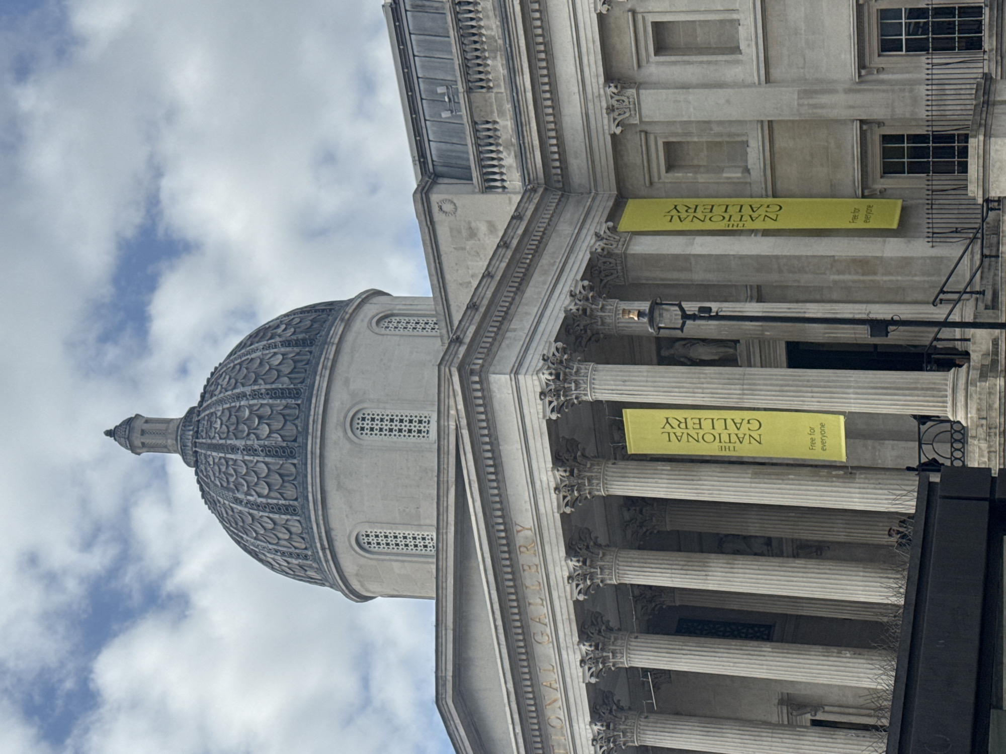 The image shows the National Gallery in London, United Kingdom, specifically highlighting the building's façade and dome against a sky with scattered clouds. The architectural features are pronounced, including the dome's patterned surface and the gallery's classical columns. Signage indicating "THE NATIONAL GALLERY" and "Free for everyone" are visible on banners.