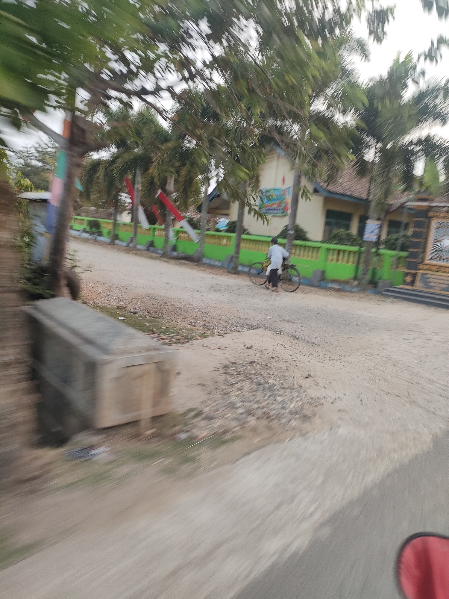 A person rides a bicycle on a dirt road in Sukoyoso, Indonesia. The road is lined with a green fence and palm trees. Behind the fence is a building with a tiled roof. The image is blurry, indicating movement.